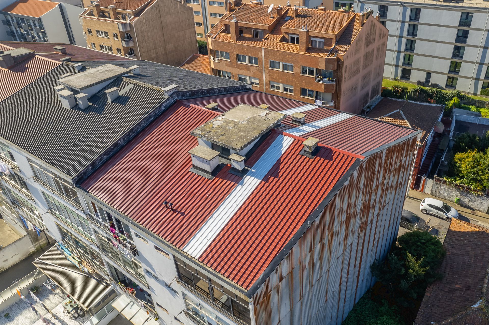 Vista de cima de prédios com telhado de metal vermelho e preto, uma faixa branca e outros prédios ao fundo.
