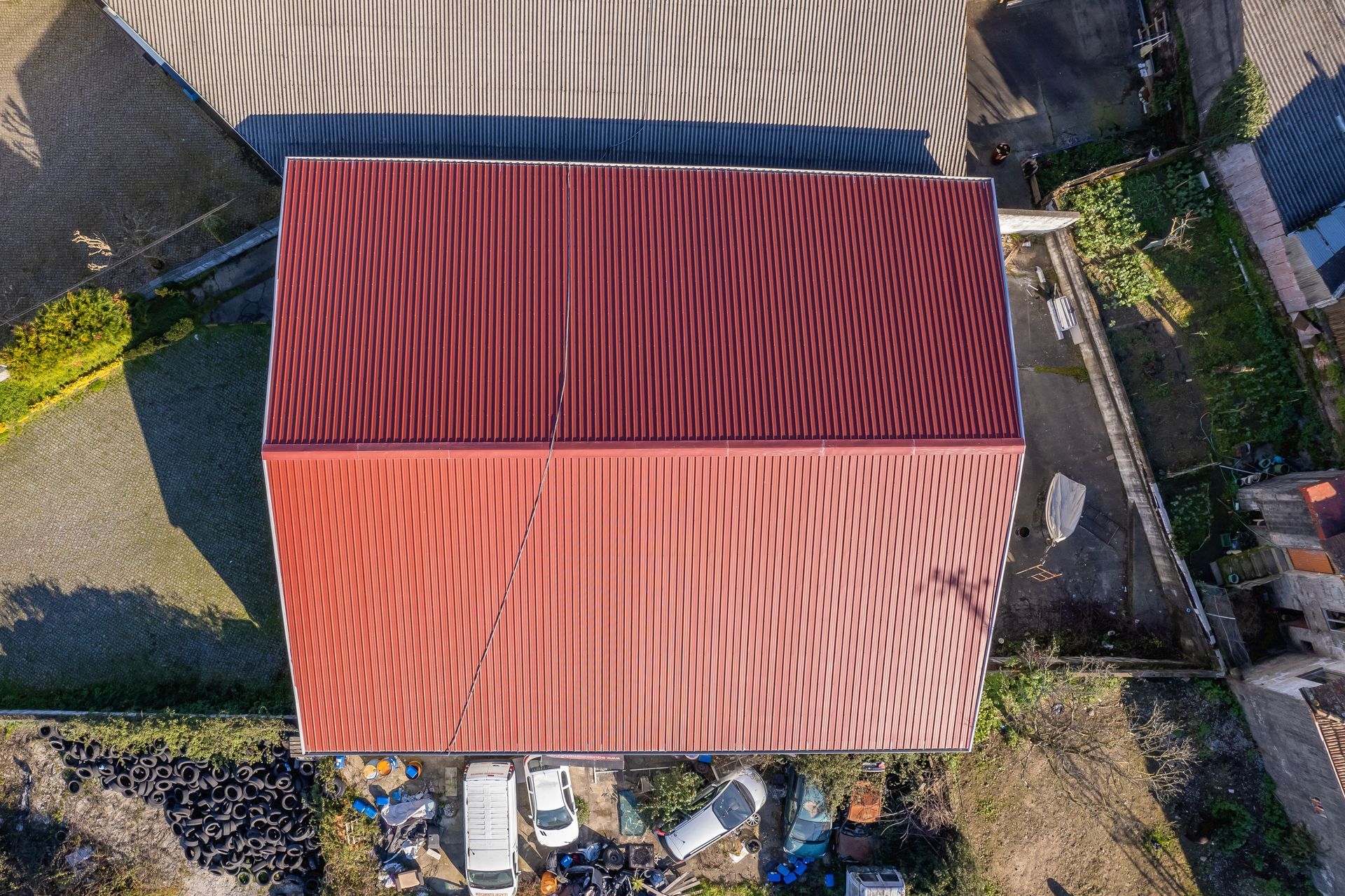 Vista aérea de um edifício com telhado de metal ondulado vermelho, carros estacionados embaixo e vegetação ao redor.