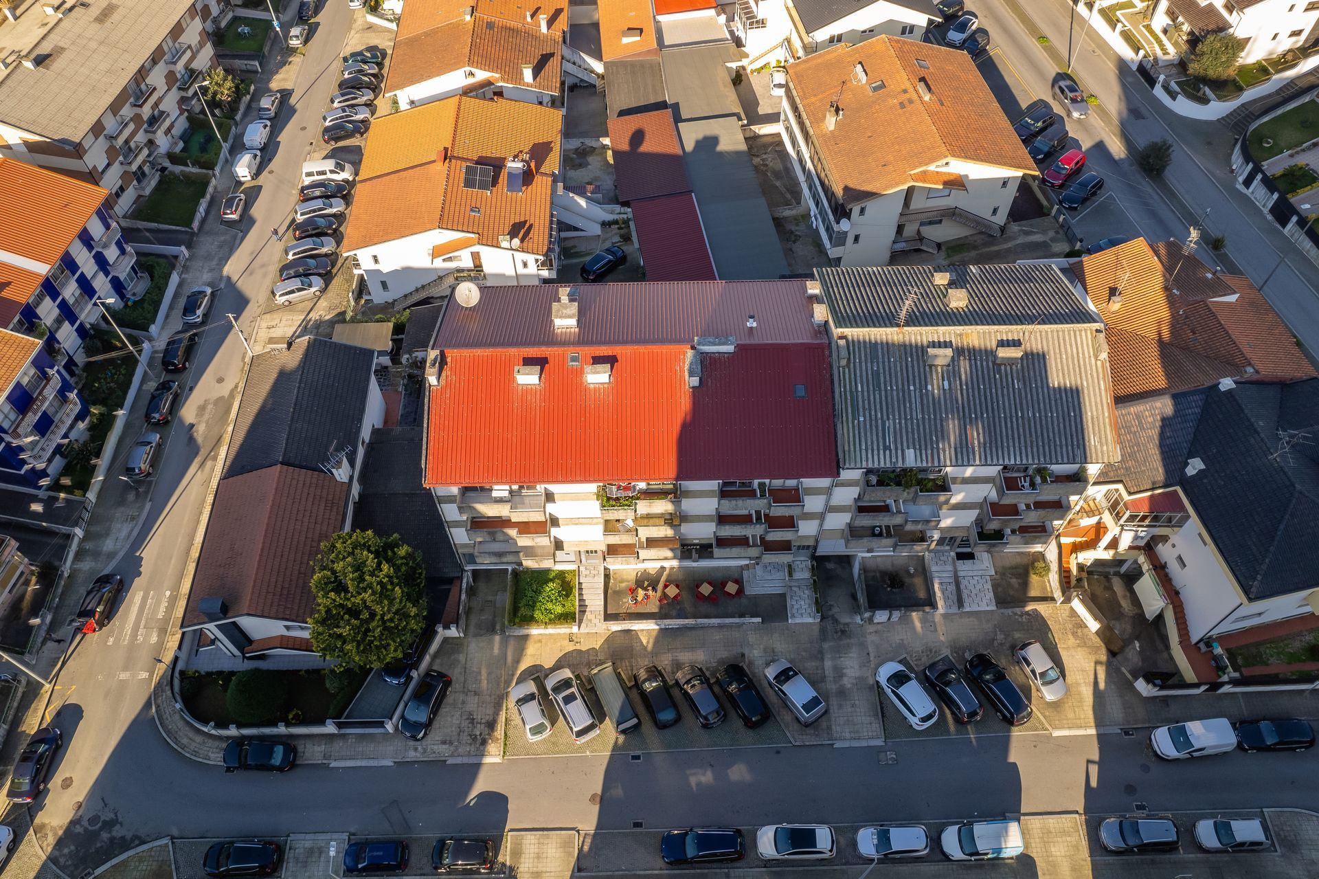 Vista aérea de prédios de apartamentos com telhados vermelhos e laranjas, cercados por carros estacionados em uma rua.
