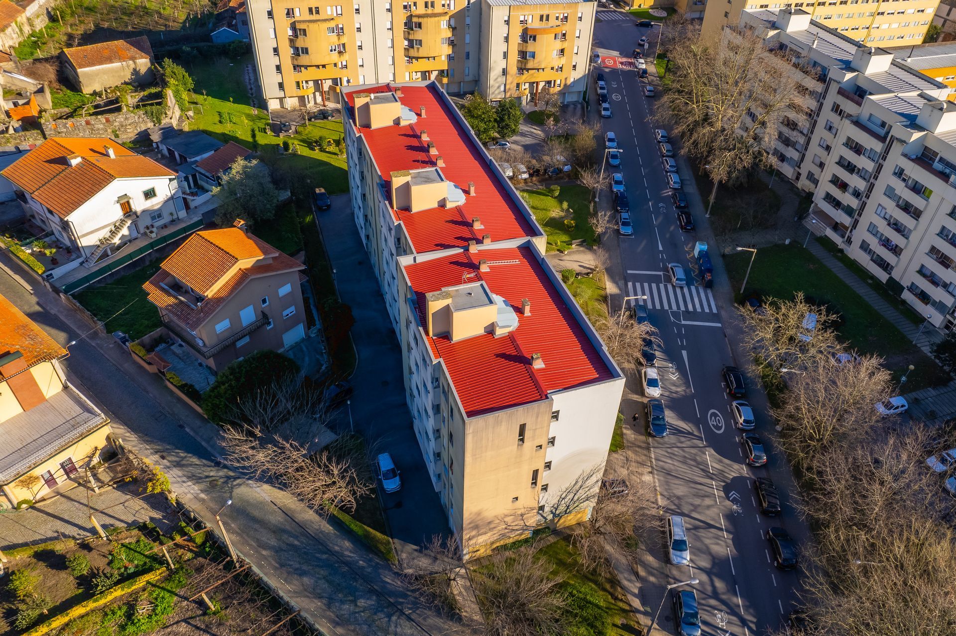 Vista aérea de um prédio comprido com telhado vermelho, situado entre ruas com carros estacionados. Outros edifícios e árvores o rodeiam.