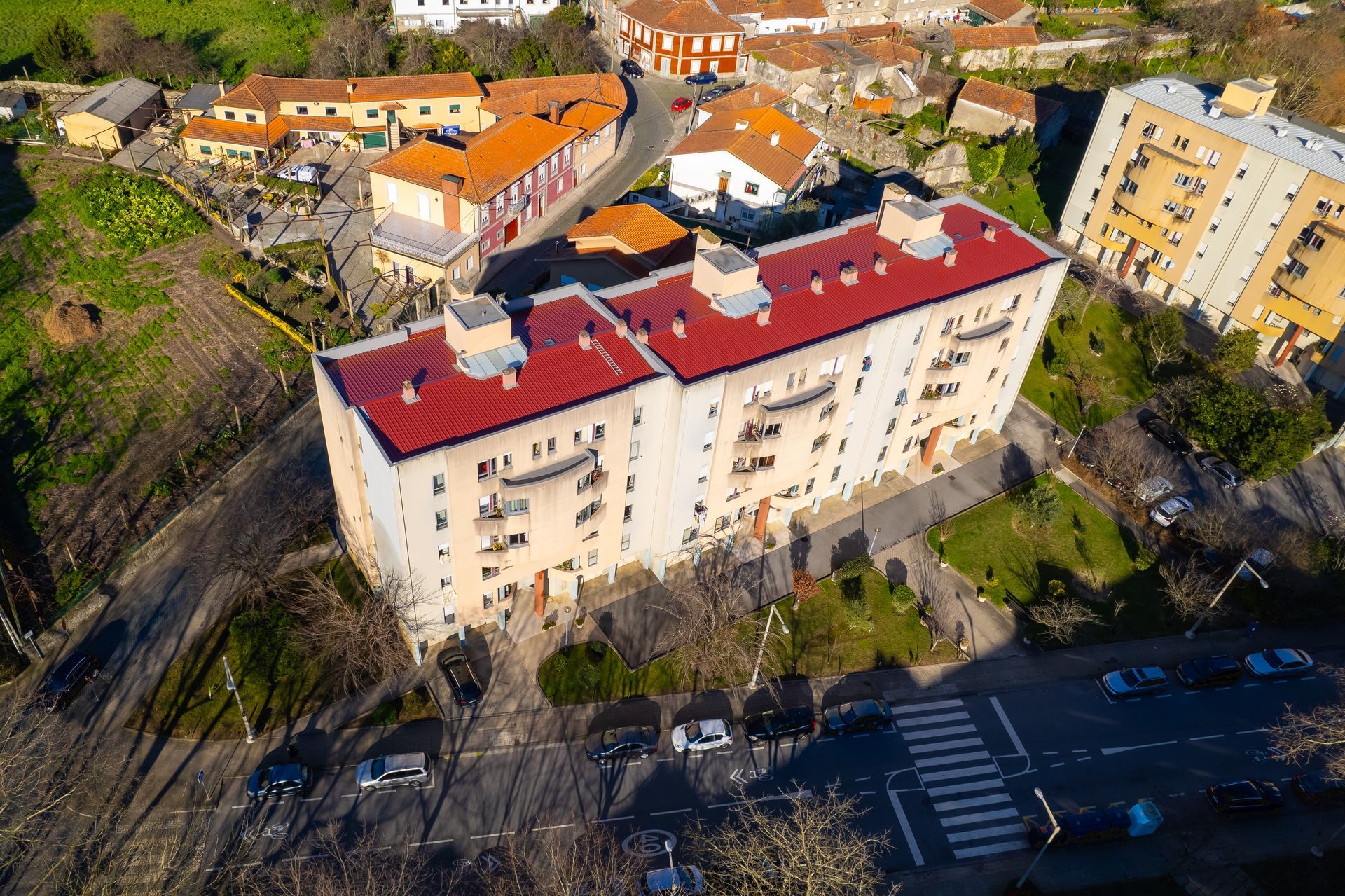 Vista aérea de um prédio de vários andares com telhado vermelho, localizado em uma cidade. A luz do sol projeta longas sombras.