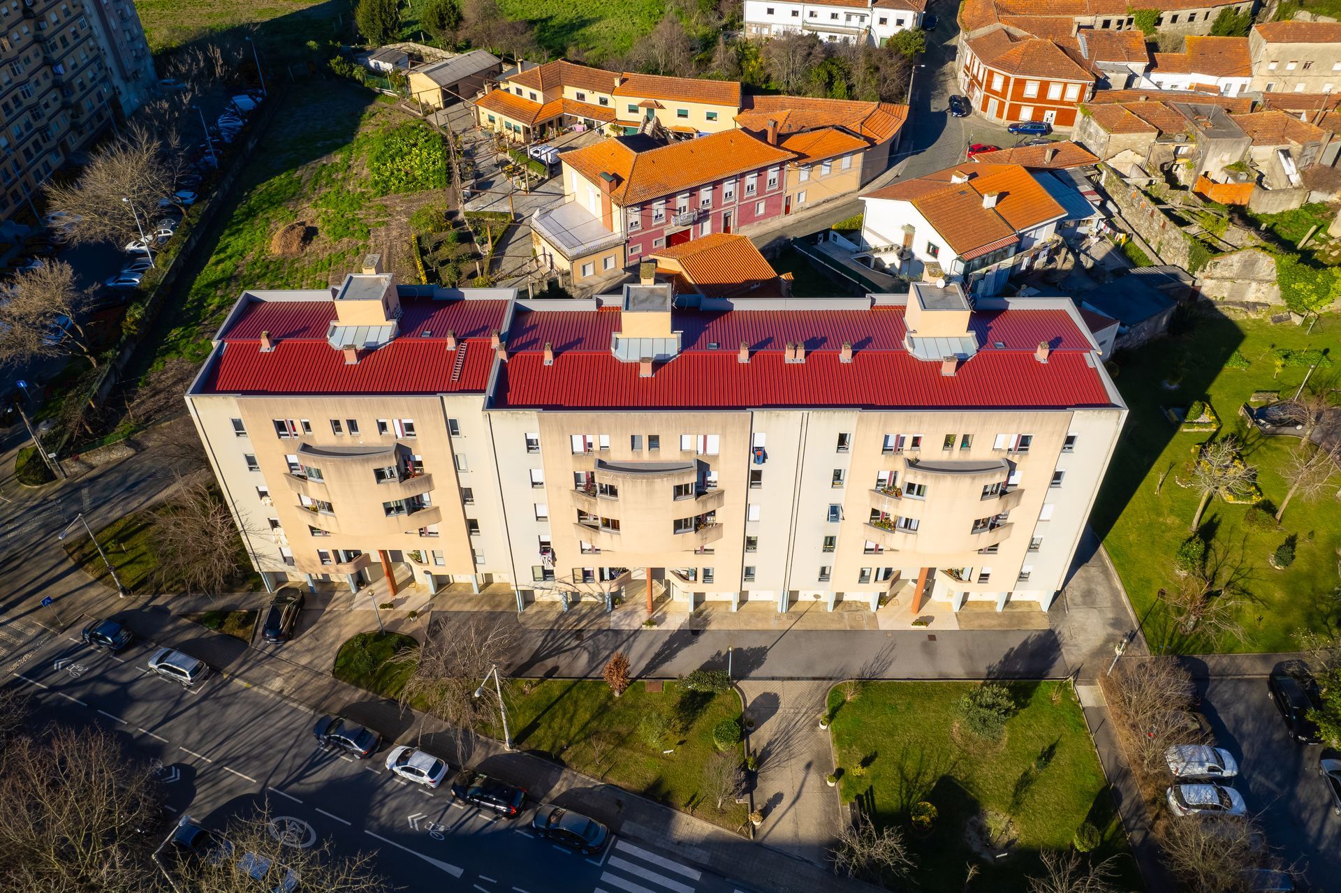 Vista aérea de um prédio de apartamentos de vários andares com telhado vermelho, localizado em uma cidade com casas de telhado laranja e grama verde.