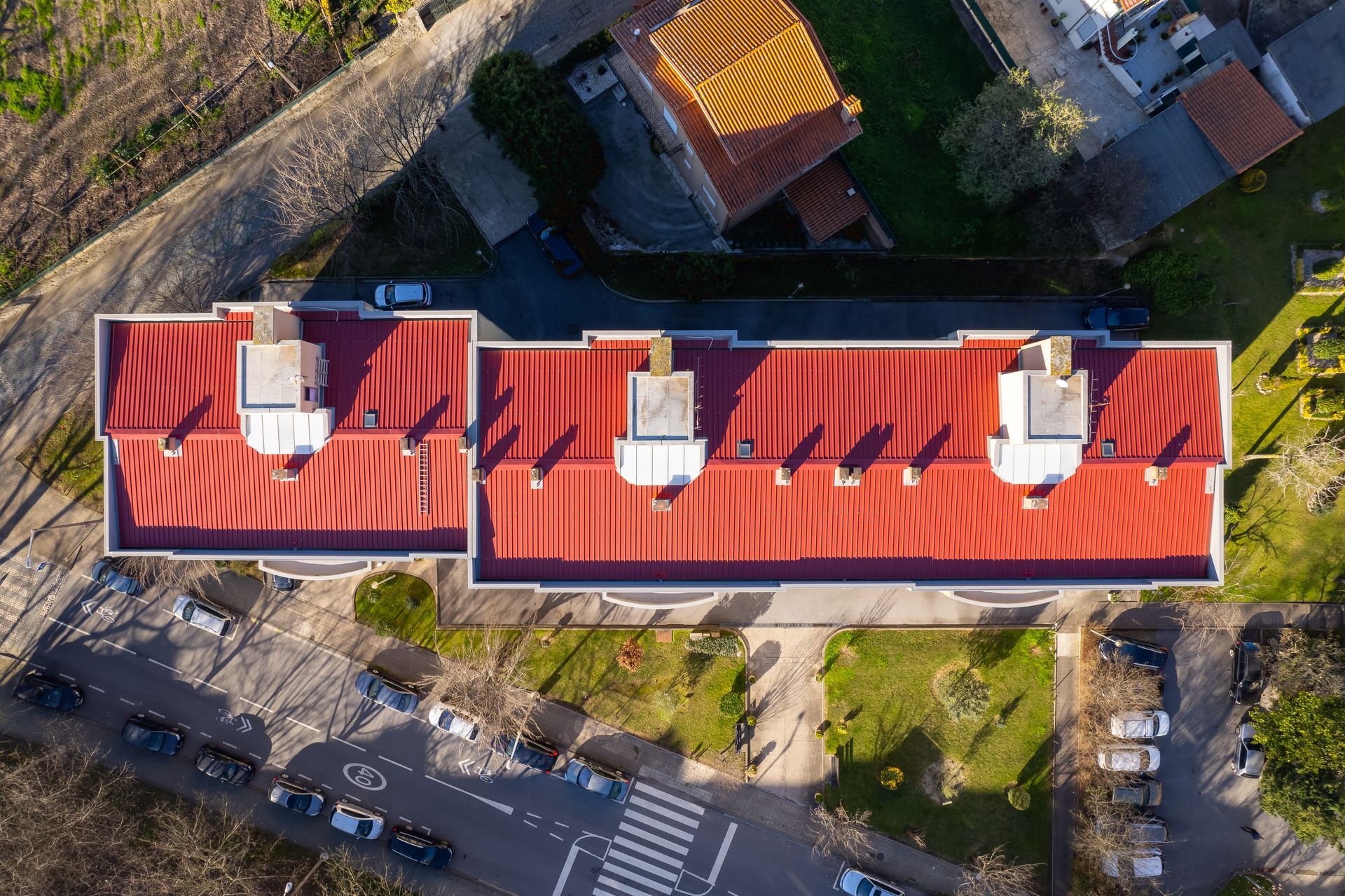 Vista aérea de um prédio com telhado vermelho projetando longas sombras em um dia ensolarado. Carros estacionados na rua.