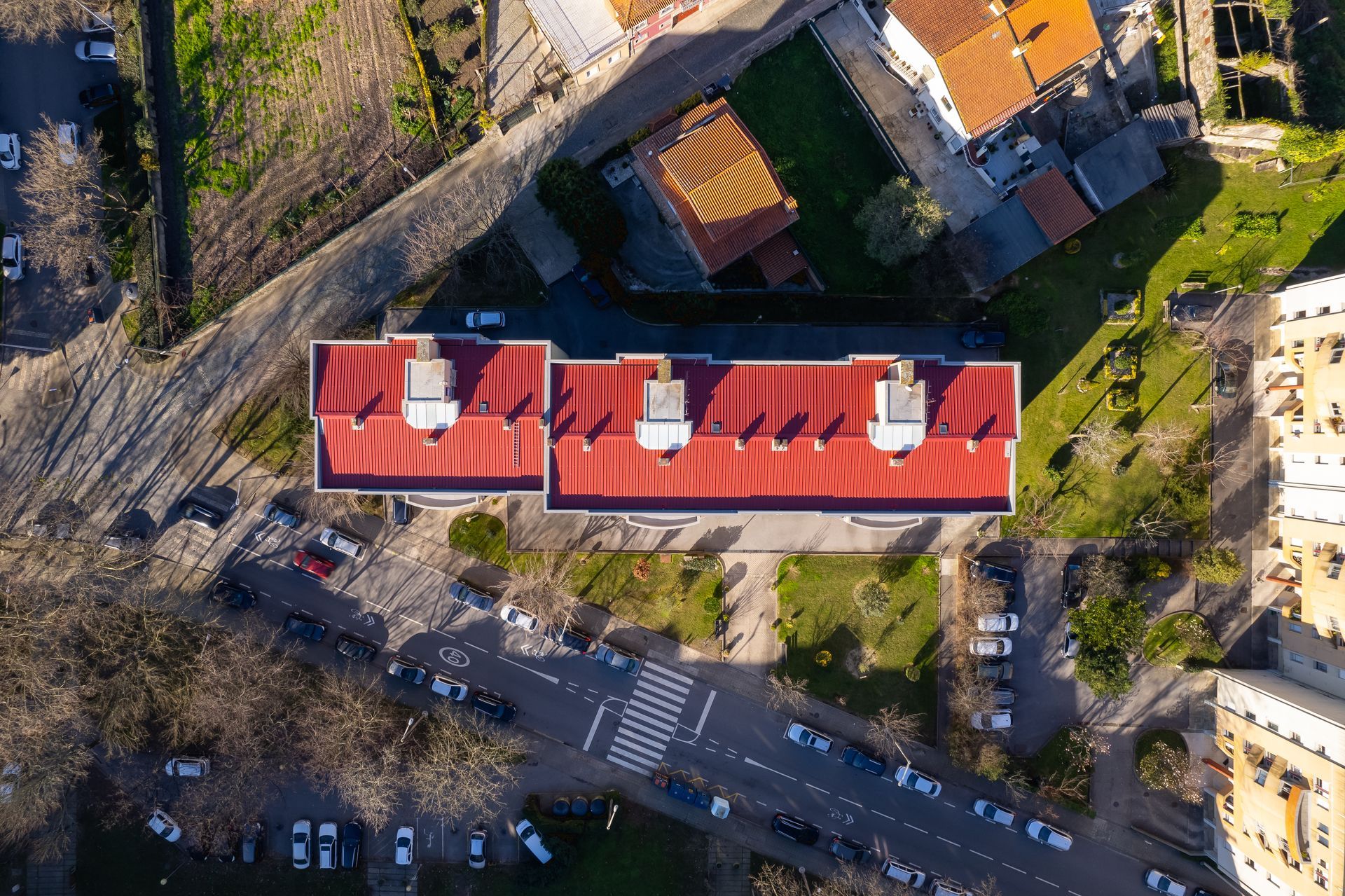 Vista aérea de um prédio com telhado de telhas vermelhas, rua e carros estacionados, com uma faixa de pedestres.