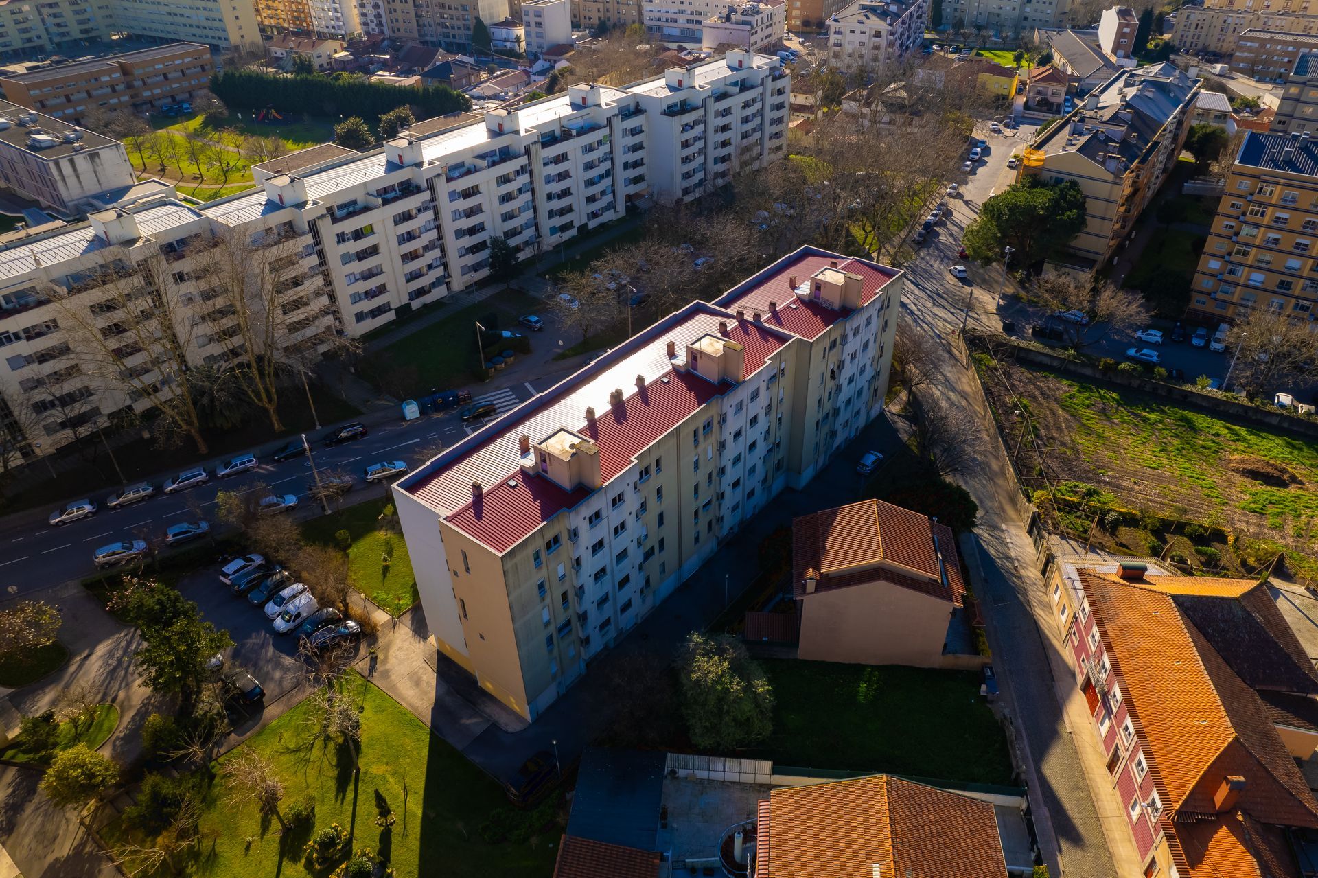 Vista aérea de edifícios de vários andares em uma cidade; dia ensolarado. Telhados bege e vermelhos, árvores, ruas com carros.