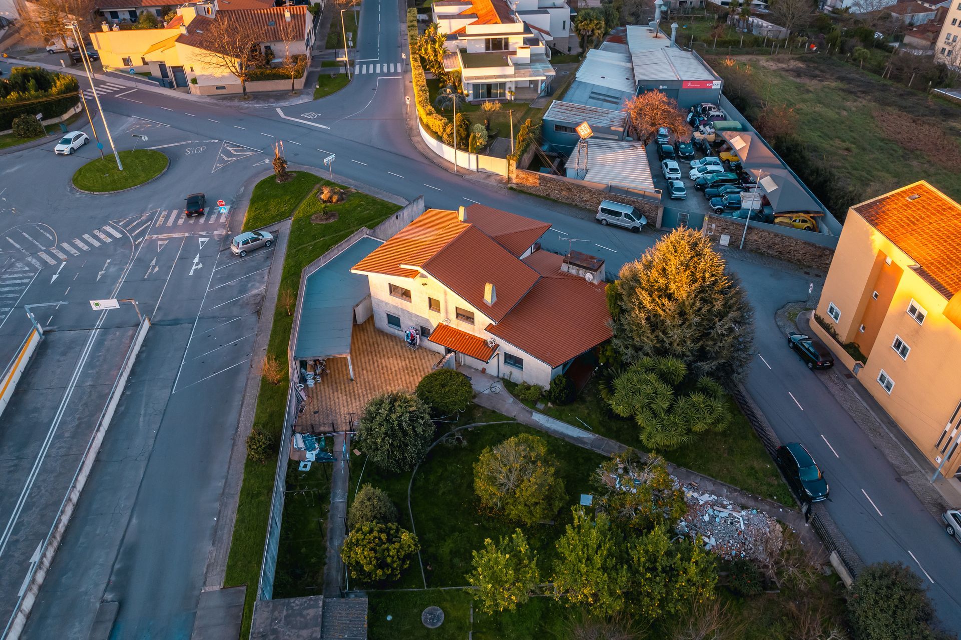 Vista aérea de uma rua com prédios, uma casa com telhado vermelho e uma rotunda, ao entardecer.