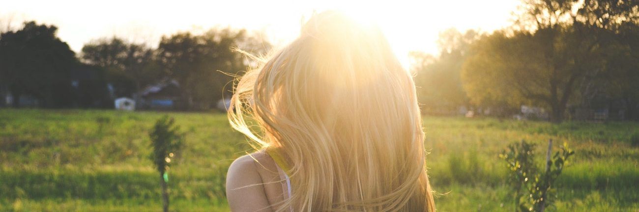 Person with long blonde hair in a sunlit grassy field at sunset