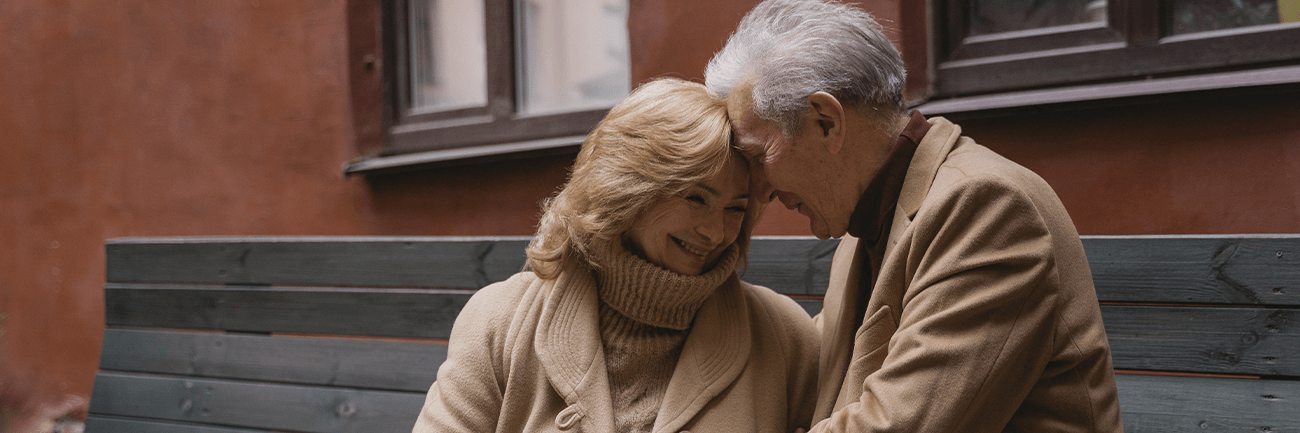 Two people sit closely on a bench, leaning together in a warm, intimate outdoor moment.