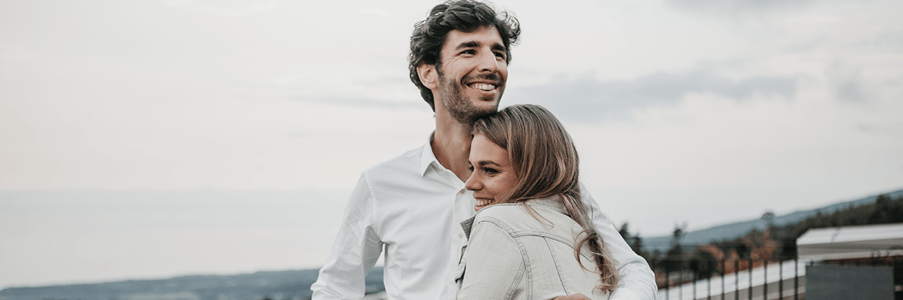 Smiling couple embracing outdoors by a waterfront under a cloudy sky