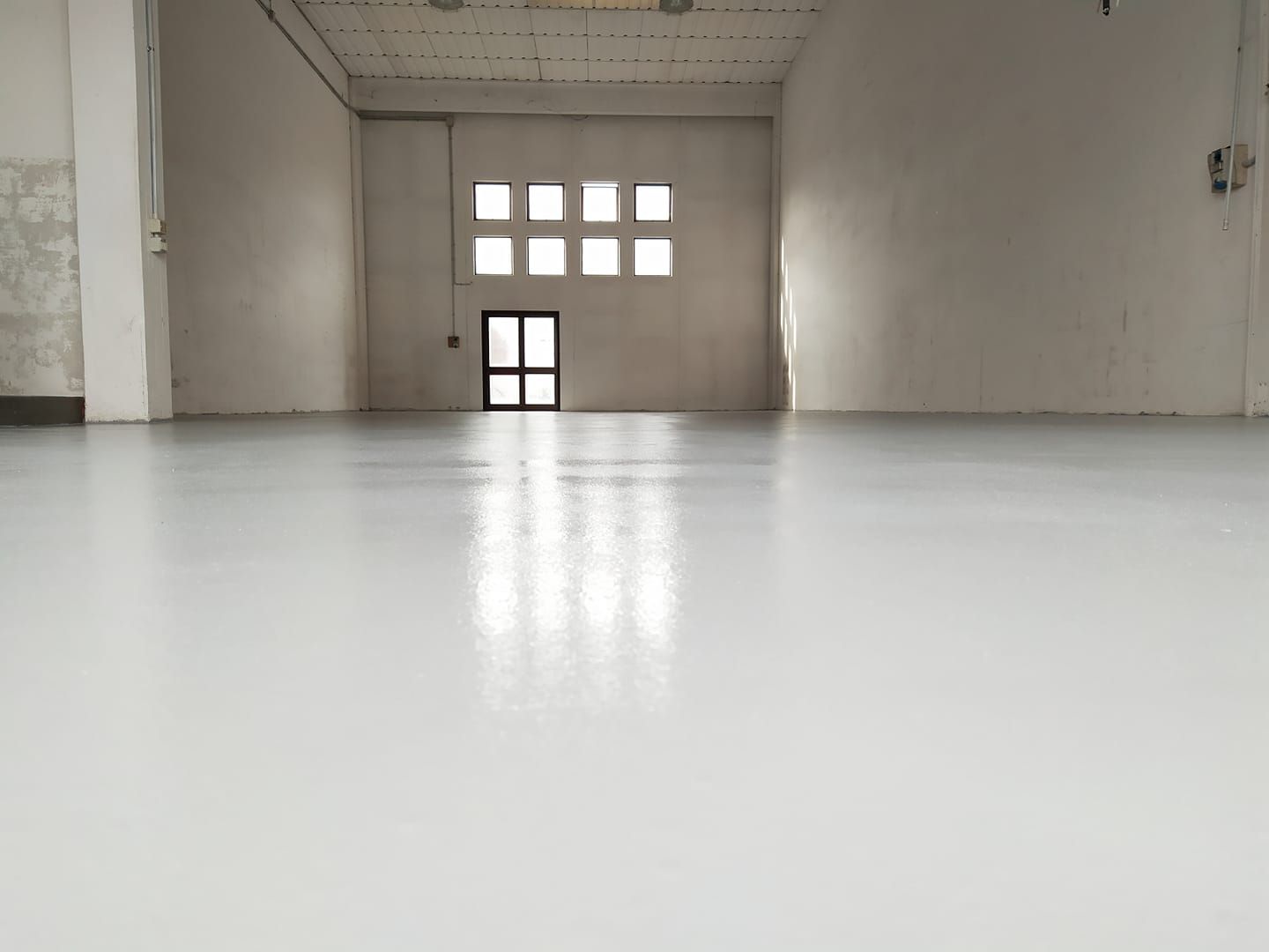 Spazio industriale vuoto con pavimento grigio chiaro lucido, finestre e una porta sul retro.