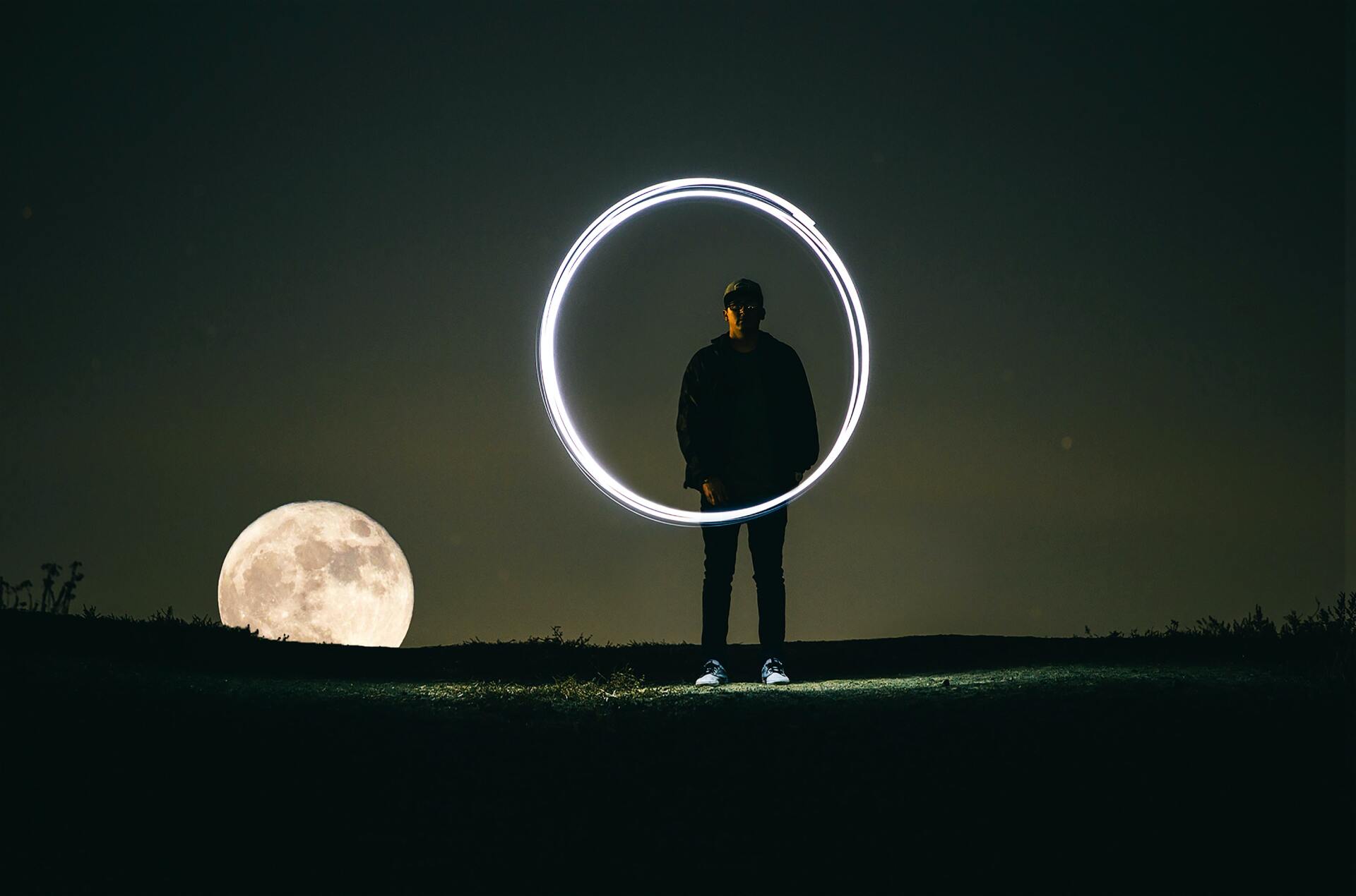 A man is standing in front of a full moon while holding a light circle.