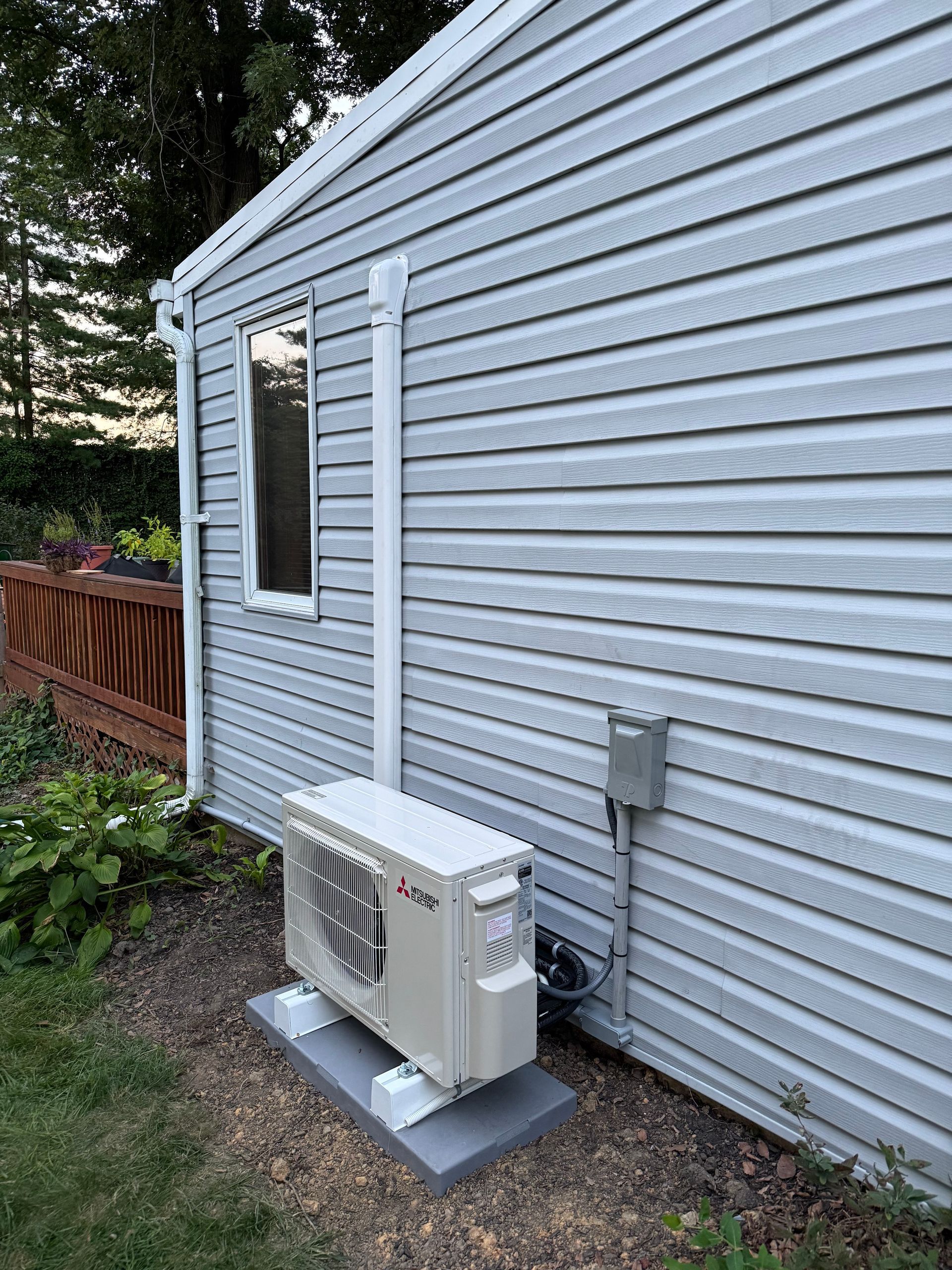 Outdoor heat pump unit on a concrete pad next to a house with gray siding and a white pipe.