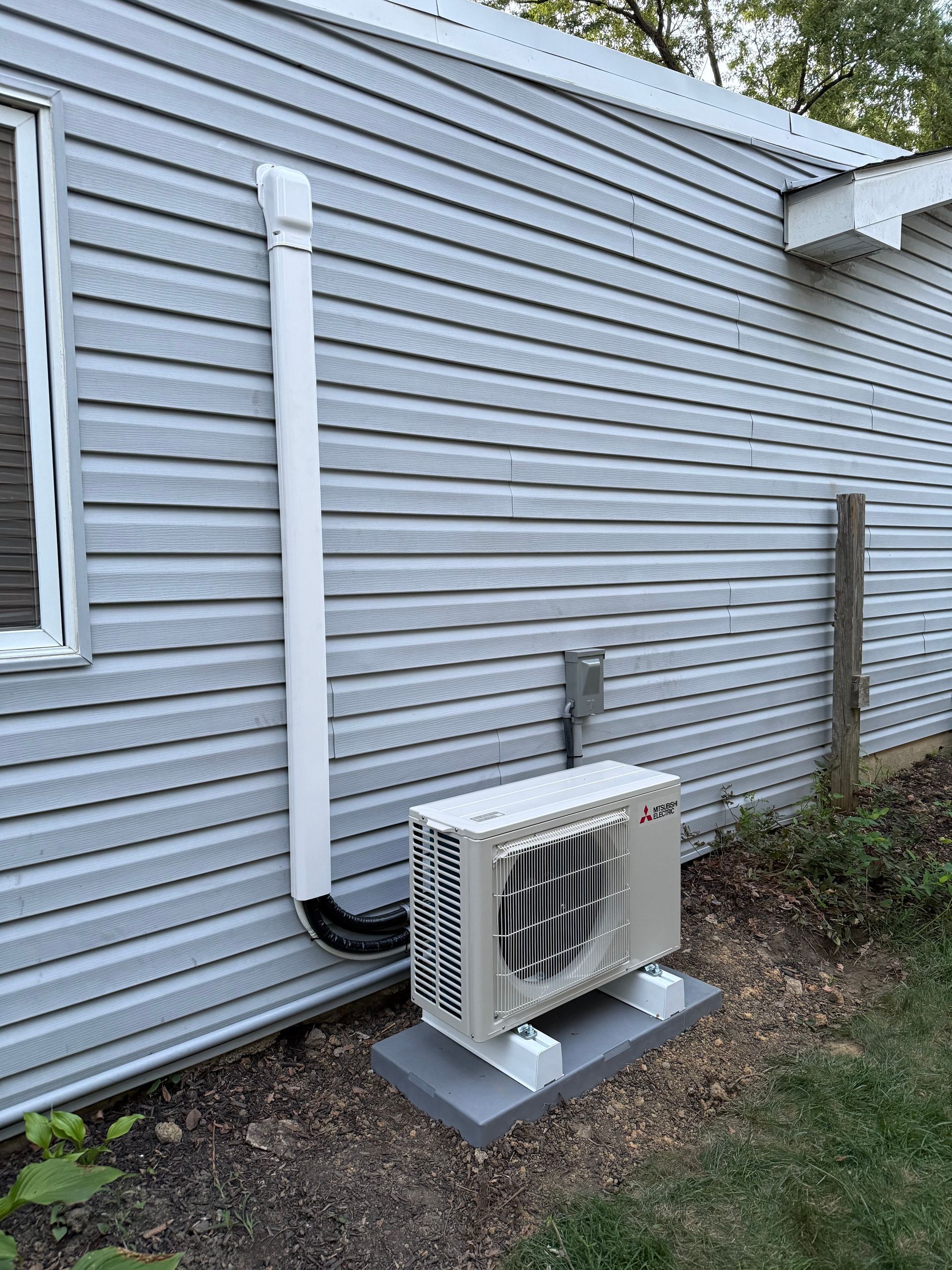 An outdoor HVAC unit on a concrete pad, against a light gray-sided building. A white exhaust pipe extends up the wall.