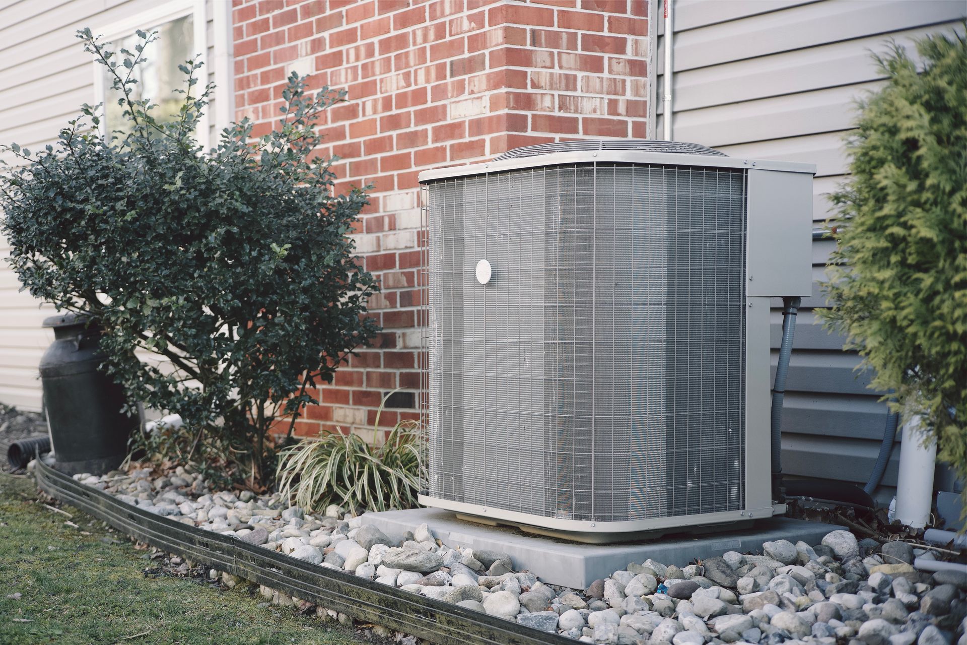 Central air conditioning unit next to brick wall and bushes on a bed of rocks.
