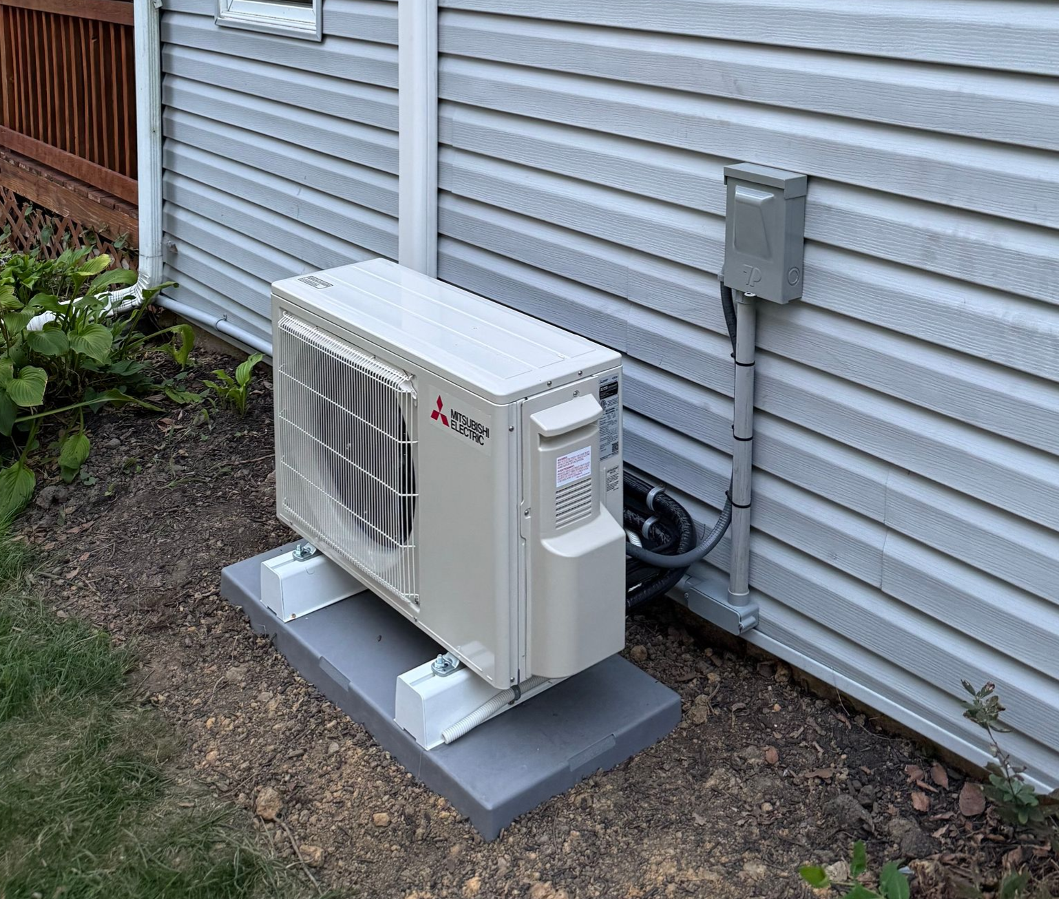 A Mitsubishi outdoor HVAC unit sits on a gray concrete pad next to the vinyl-sided wall of a house.