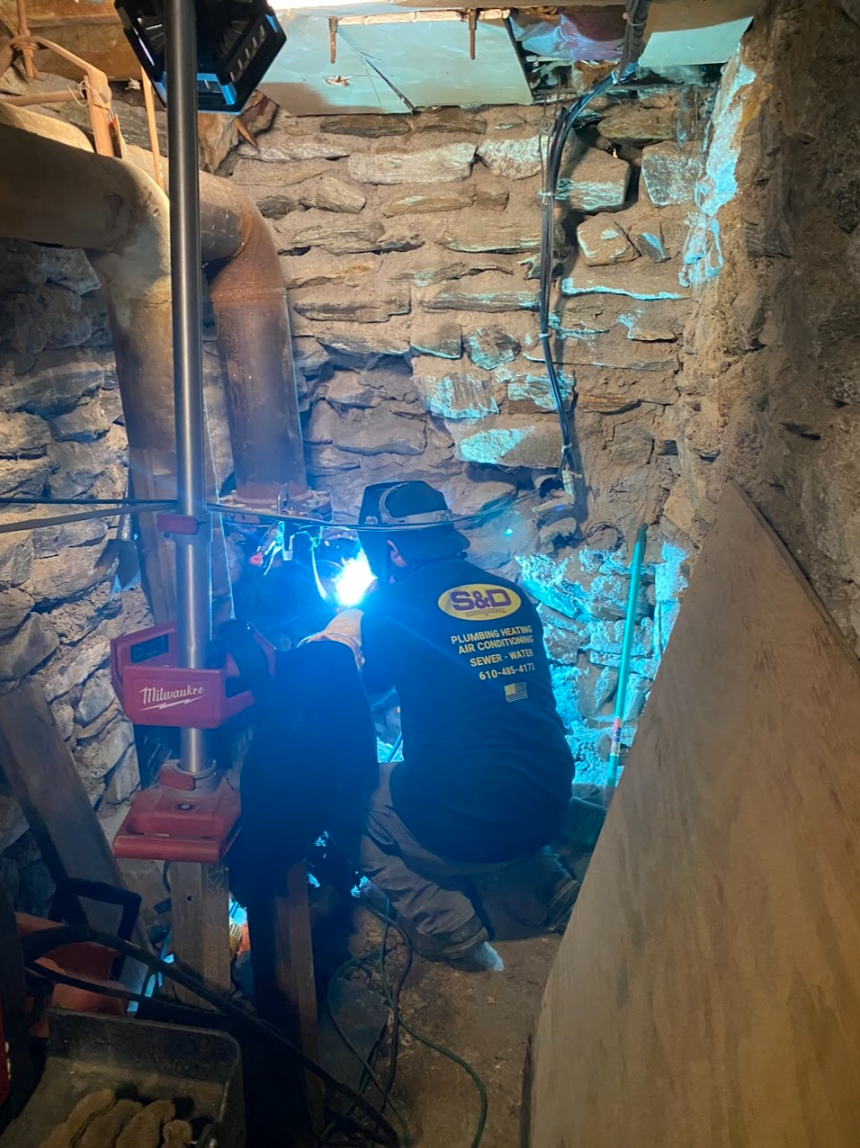 A person in dark clothing welds pipes in a dimly lit, unfinished stone basement, producing a bright blue arc.