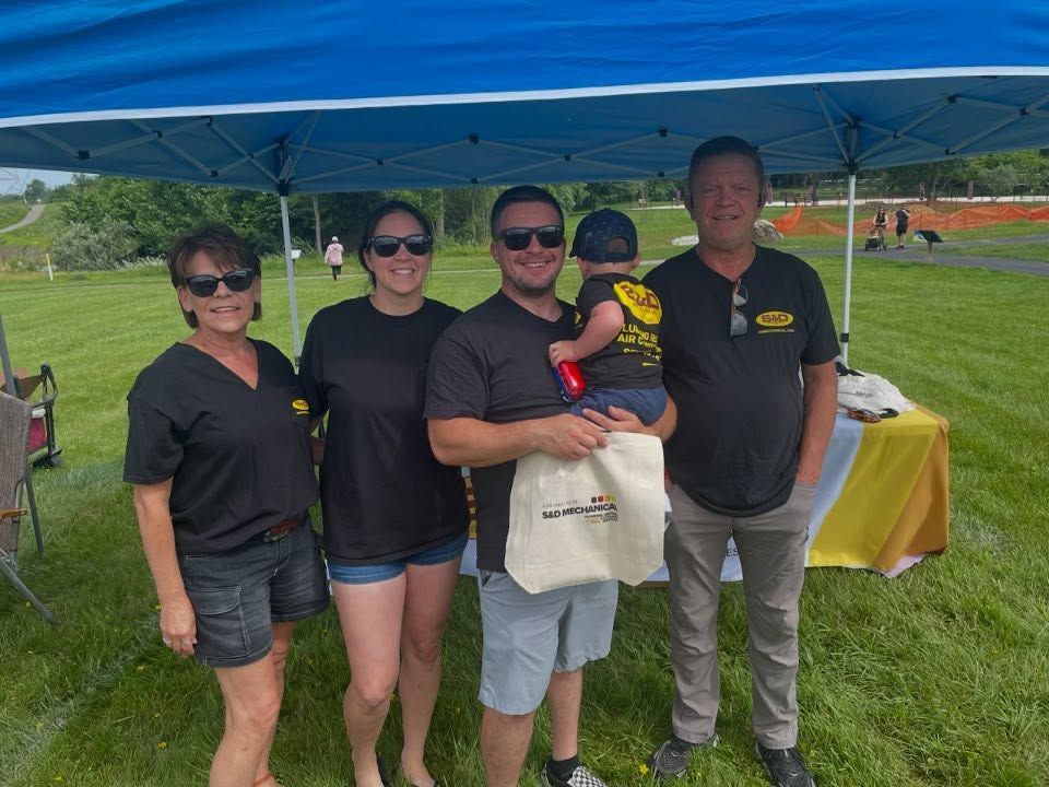 Five people pose under a blue canopy on a grassy field. One holds a baby. Table with items in front.