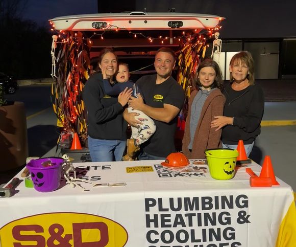 Family at a trunk-or-treat event with a plumbing company display. People smiling at a table with buckets and a hard hat.