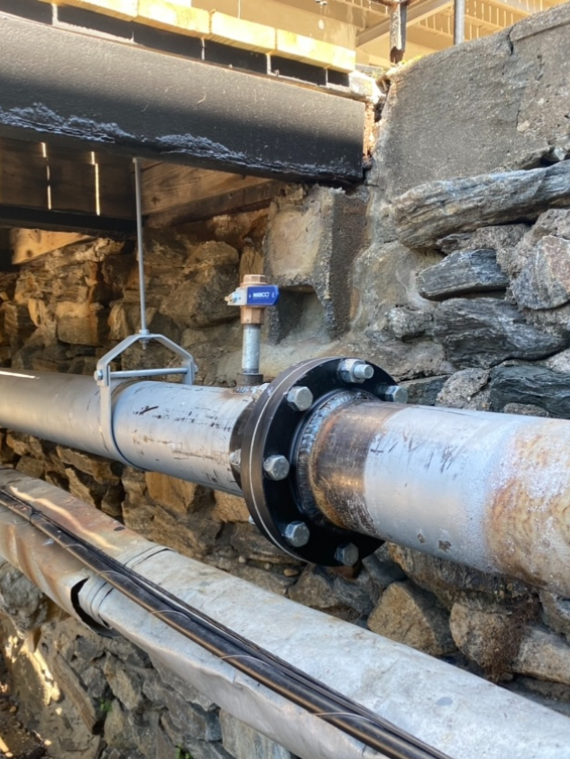 A horizontal metal pipe with a black flanged connection and a blue-handled valve, suspended against a stone foundation wall.