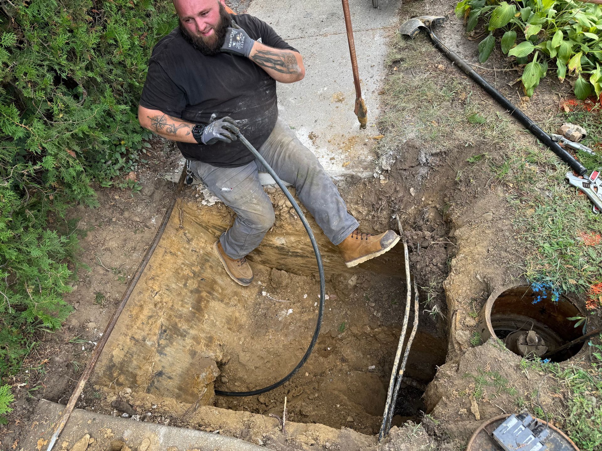 Man pulling a black hose from a hole he dug.  He wears work gloves, sits in the hole, and holds the hose.