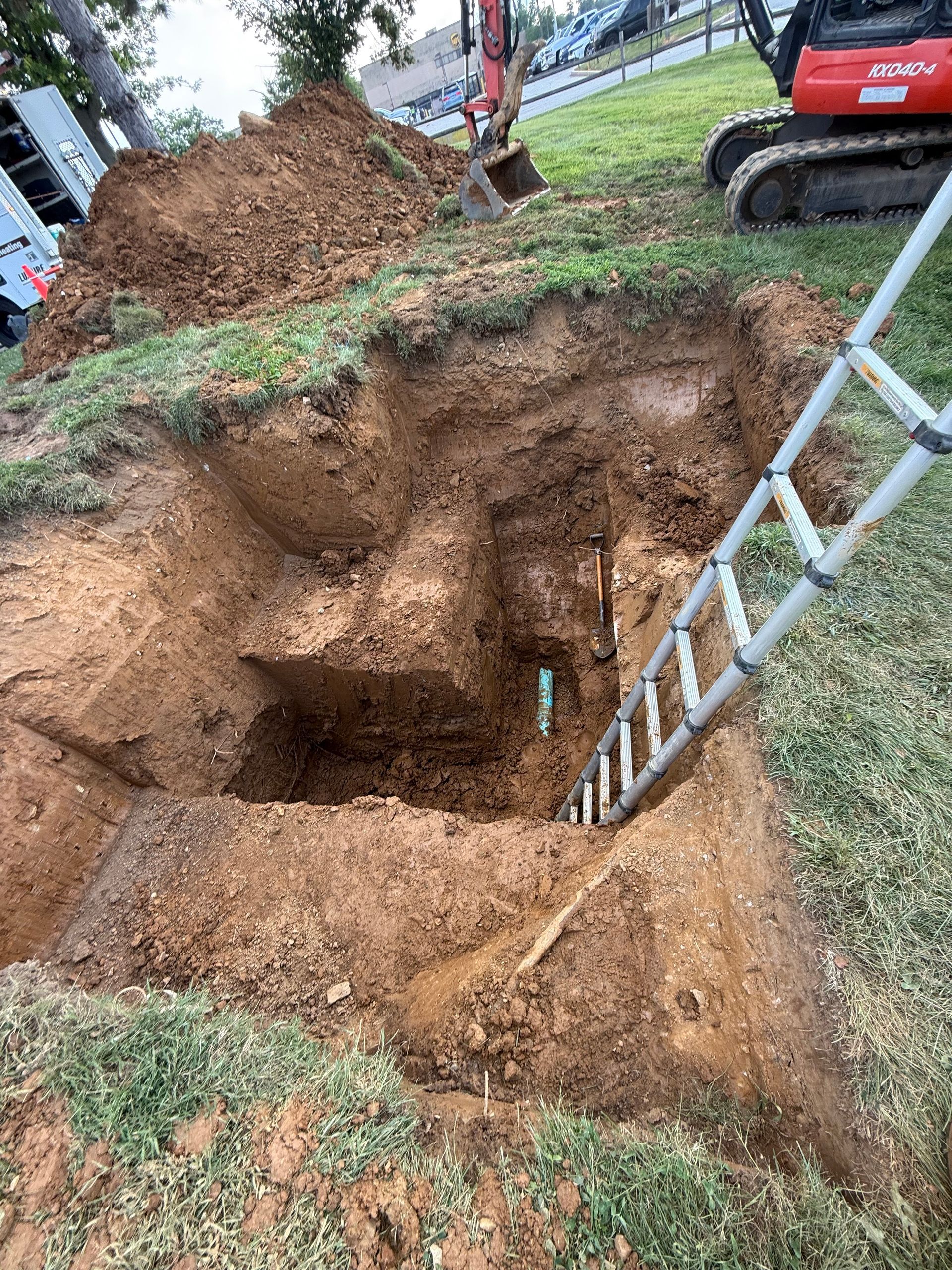 Large square excavation in a grassy area with ladder, mini-excavator, and dirt pile; likely construction.