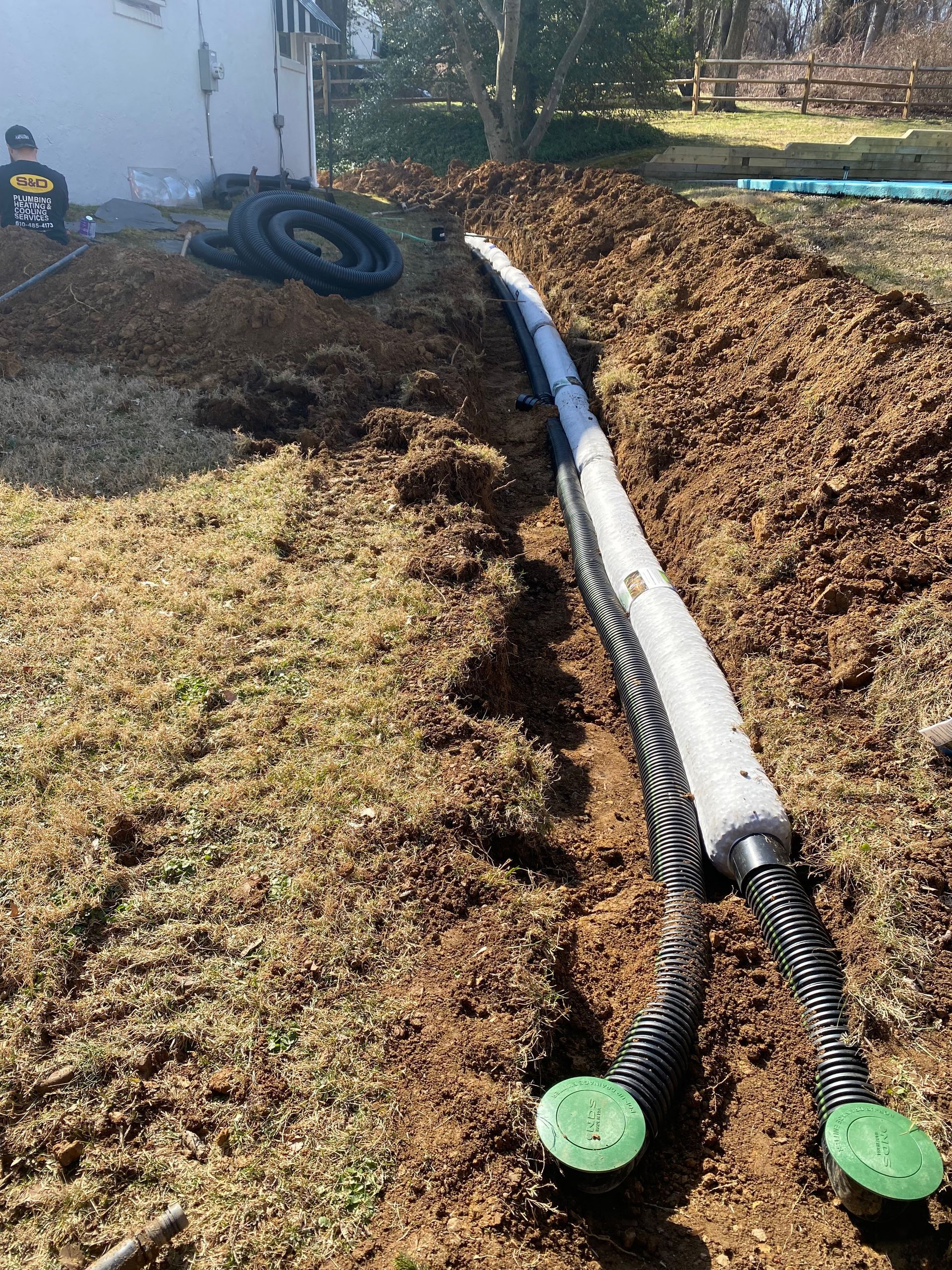 A trench with drainage pipe installed in a yard. Ground is brown dirt and dry grass.