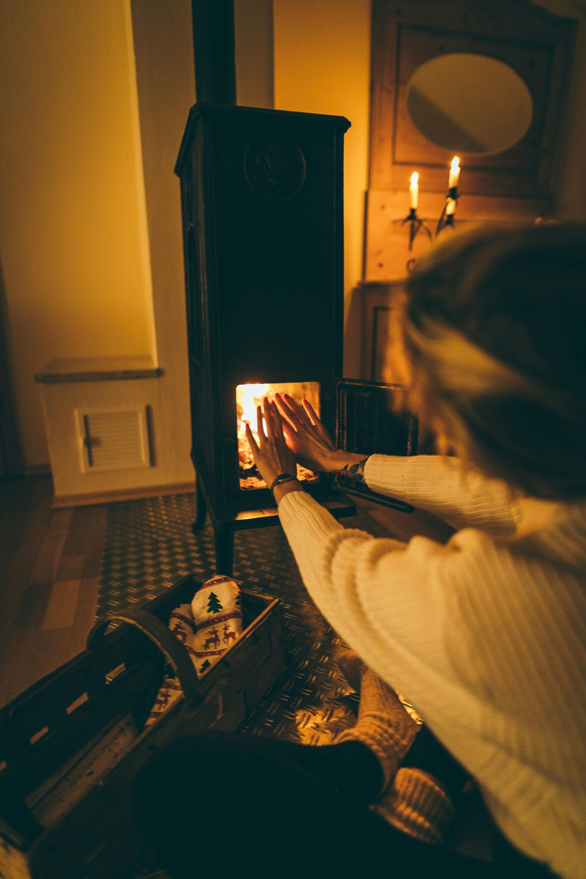 Woman warming hands by a lit wood-burning stove in a dimly lit room with candles and a basket.