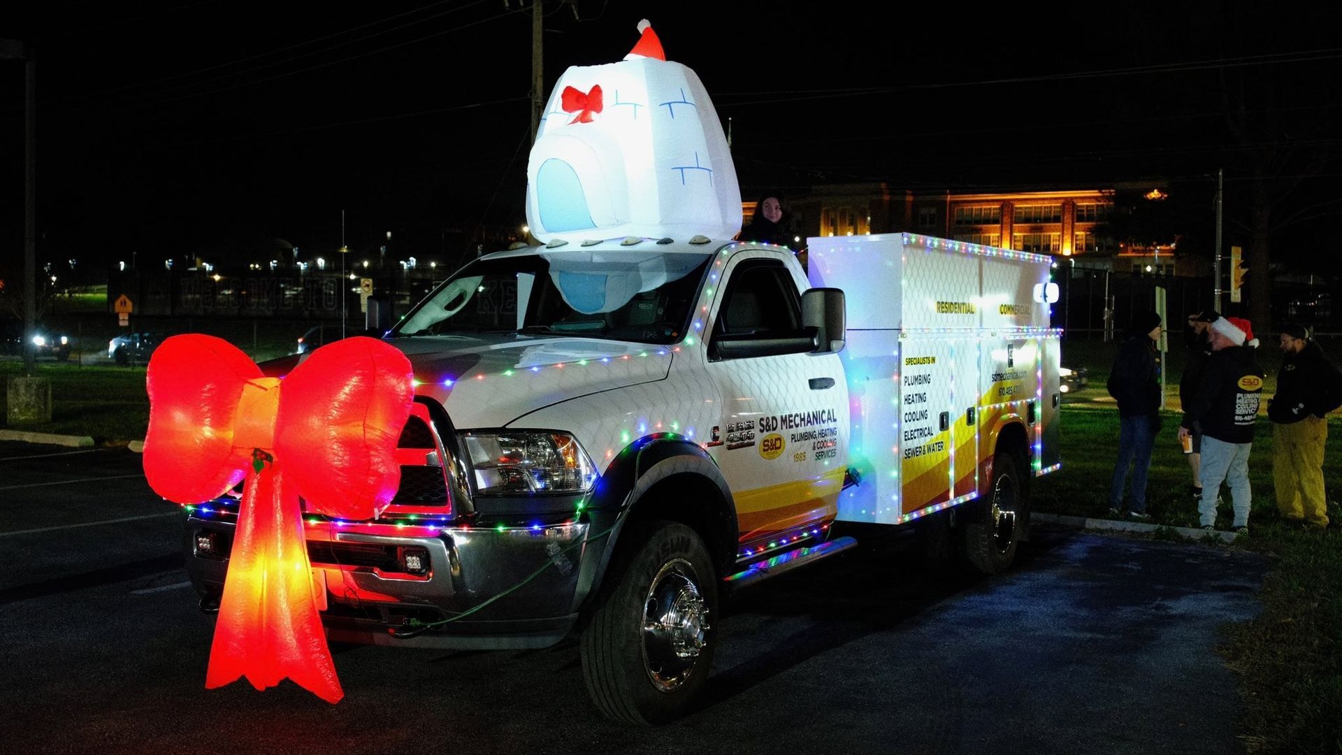 White truck decorated with lights and a large red bow, topped with an illuminated chicken for a holiday parade.