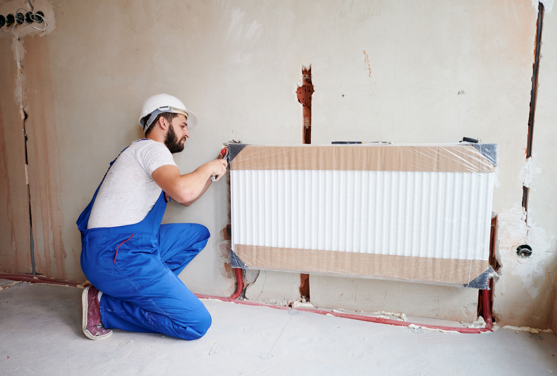 Construction worker installing a radiator on a wall, wearing blue overalls and a white hard hat.