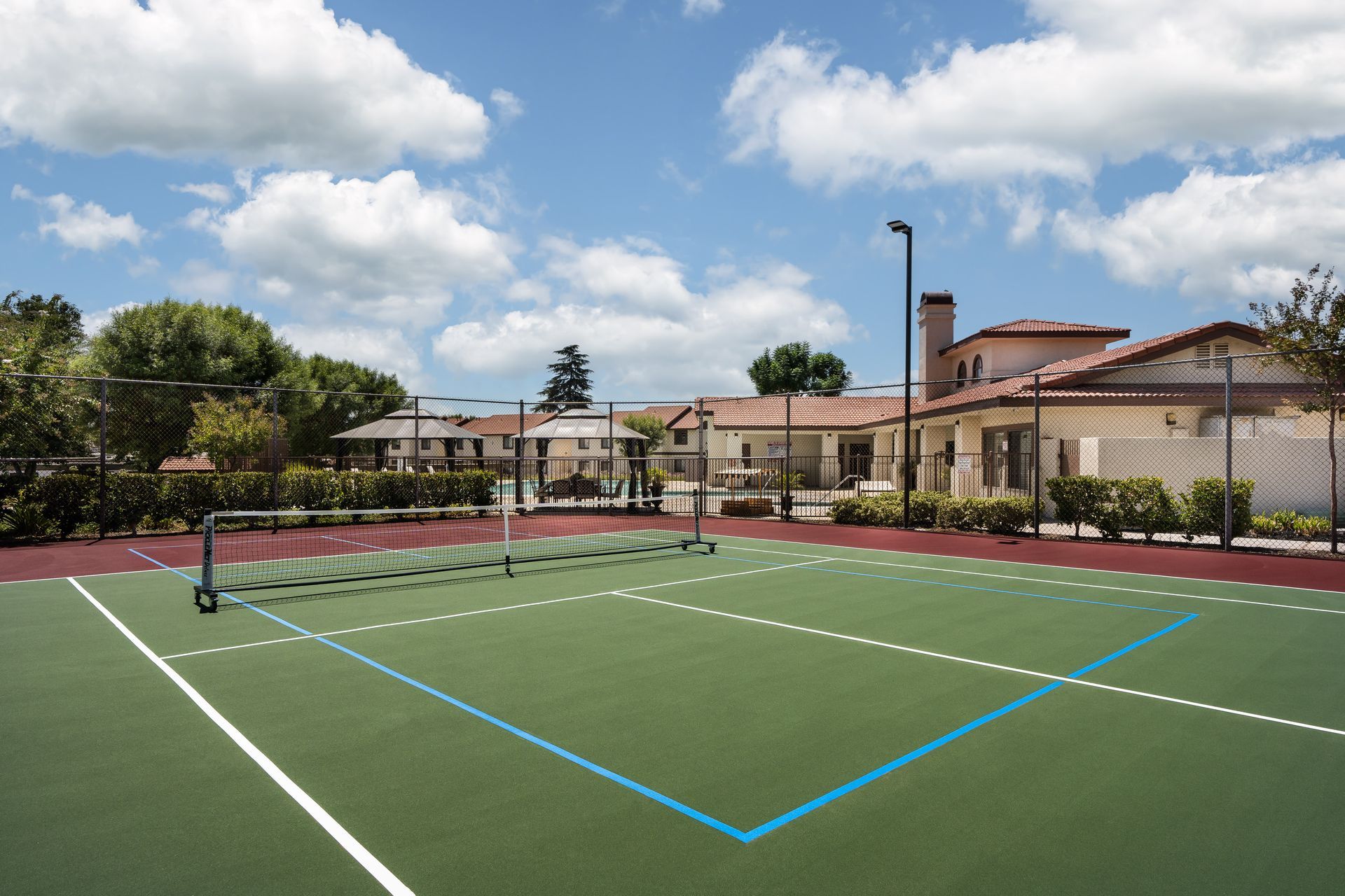 Tennis court with net, green surface, sunny day, and buildings in the background.