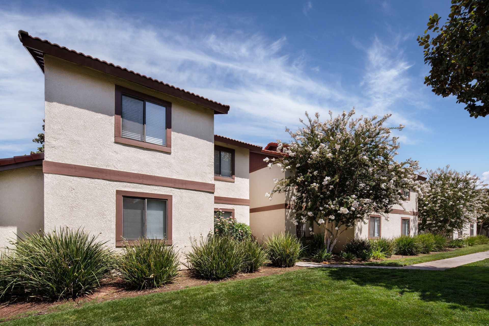 Two-story beige stucco apartments with brown trim and green lawn on a sunny day with blue sky.