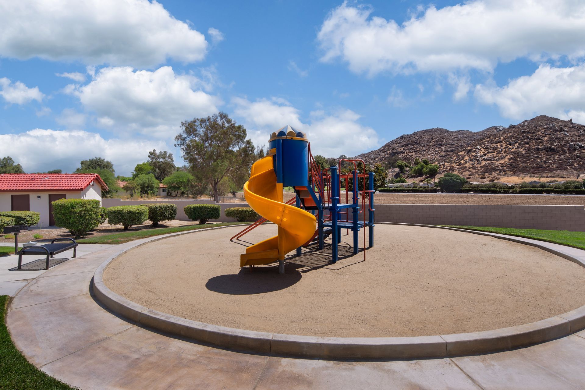 Playground with yellow slide, blue supports, and tan sand. In a park setting with mountains in the background.