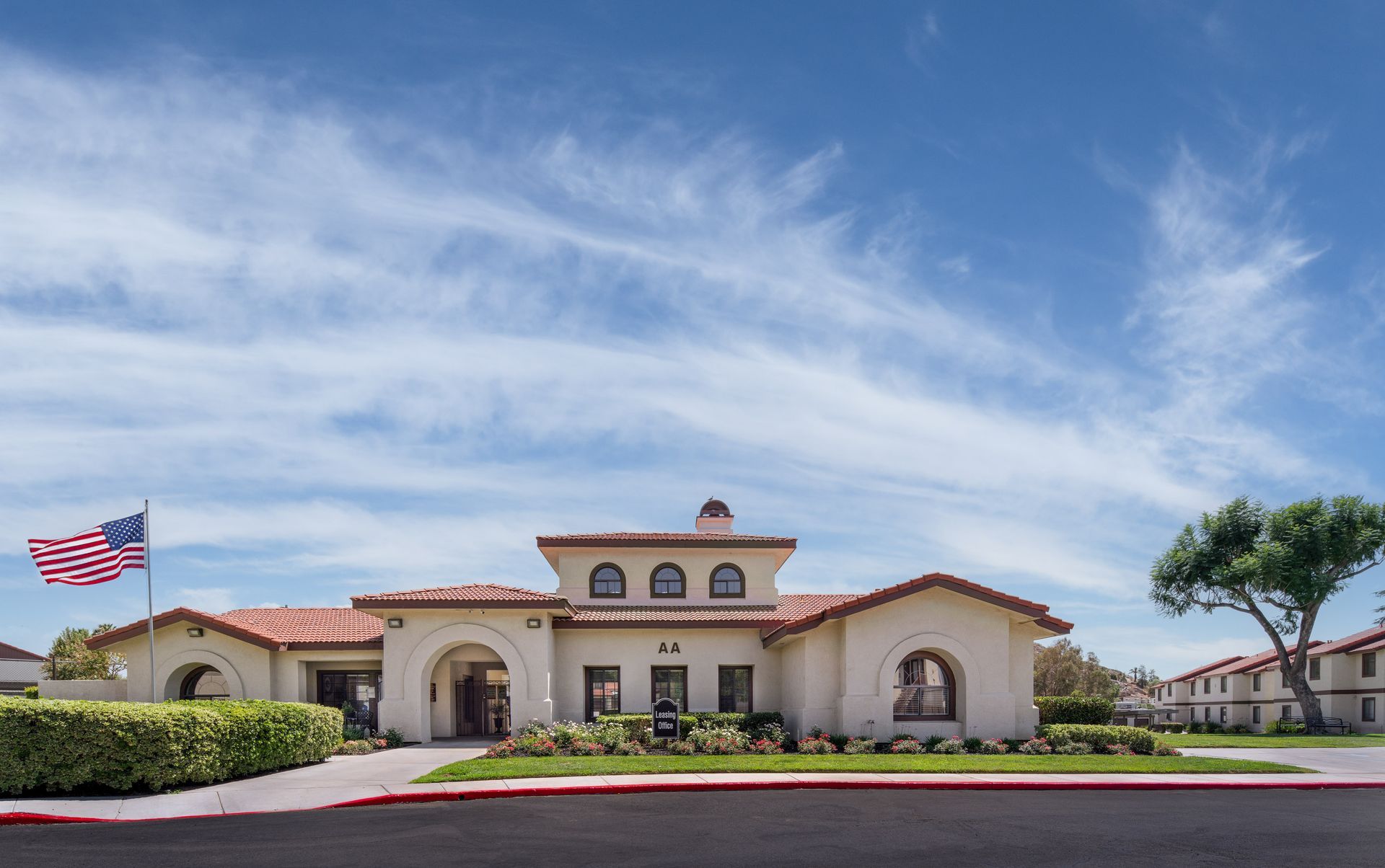 A light-colored building with a red tile roof, American flag, and cloudy blue sky.