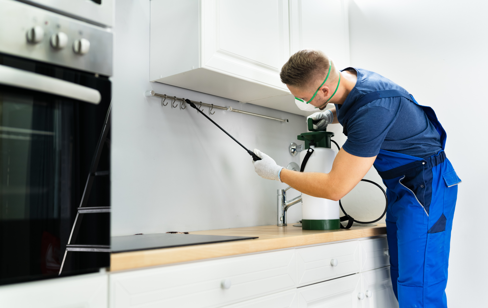 Technician spraying cleaner under a white kitchen cabinet beside a stove
