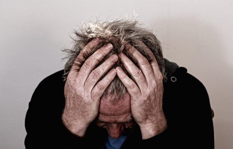 Person with gray hair sits hunched, covering their face with both hands against a plain background