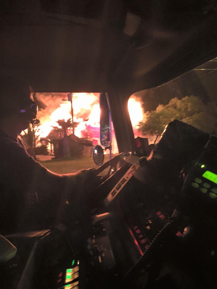 A man is driving a fire truck with a fire in the background