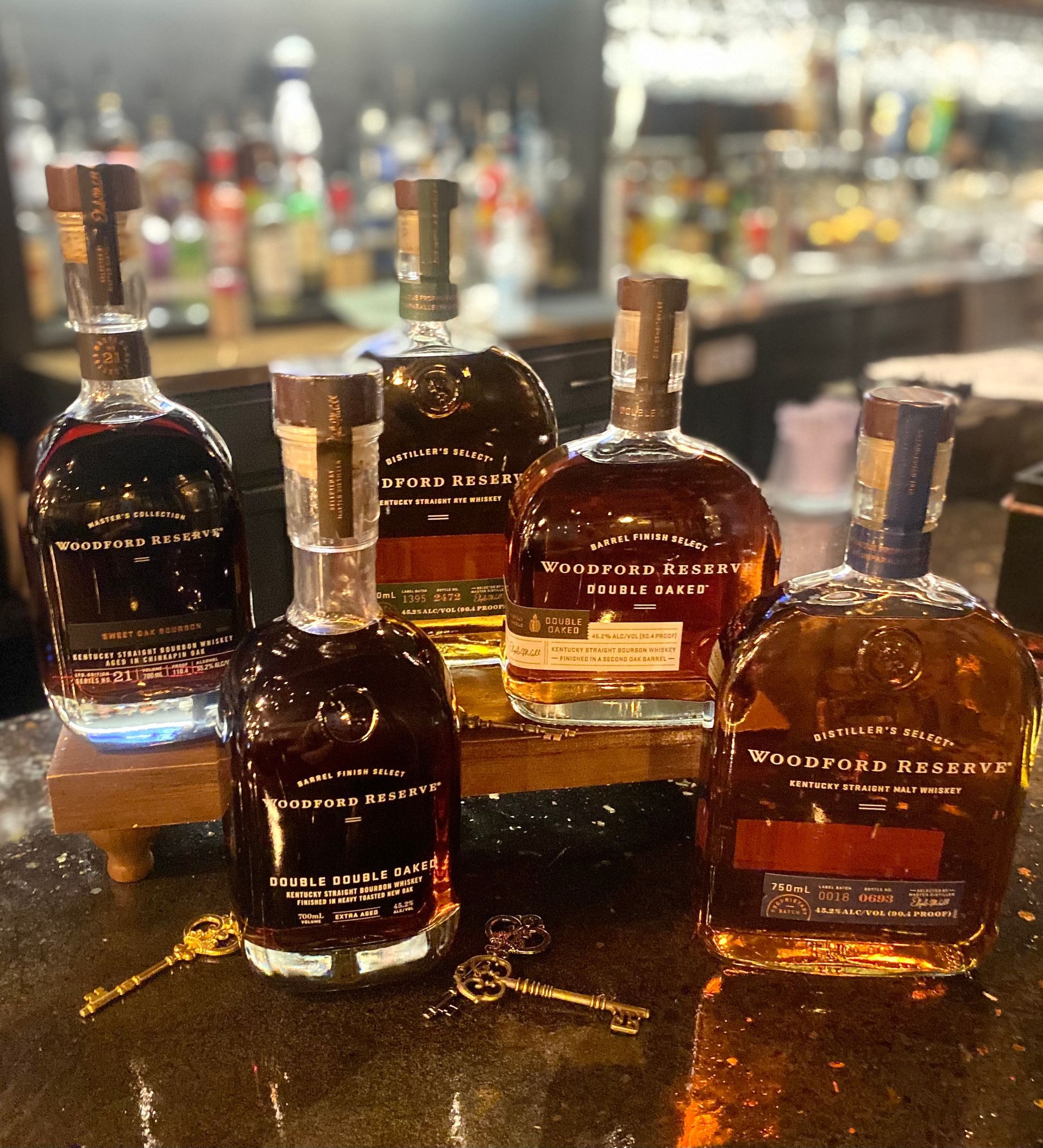 Several bottles of bourbon arranged on a bar counter with a blurred liquor display in the background