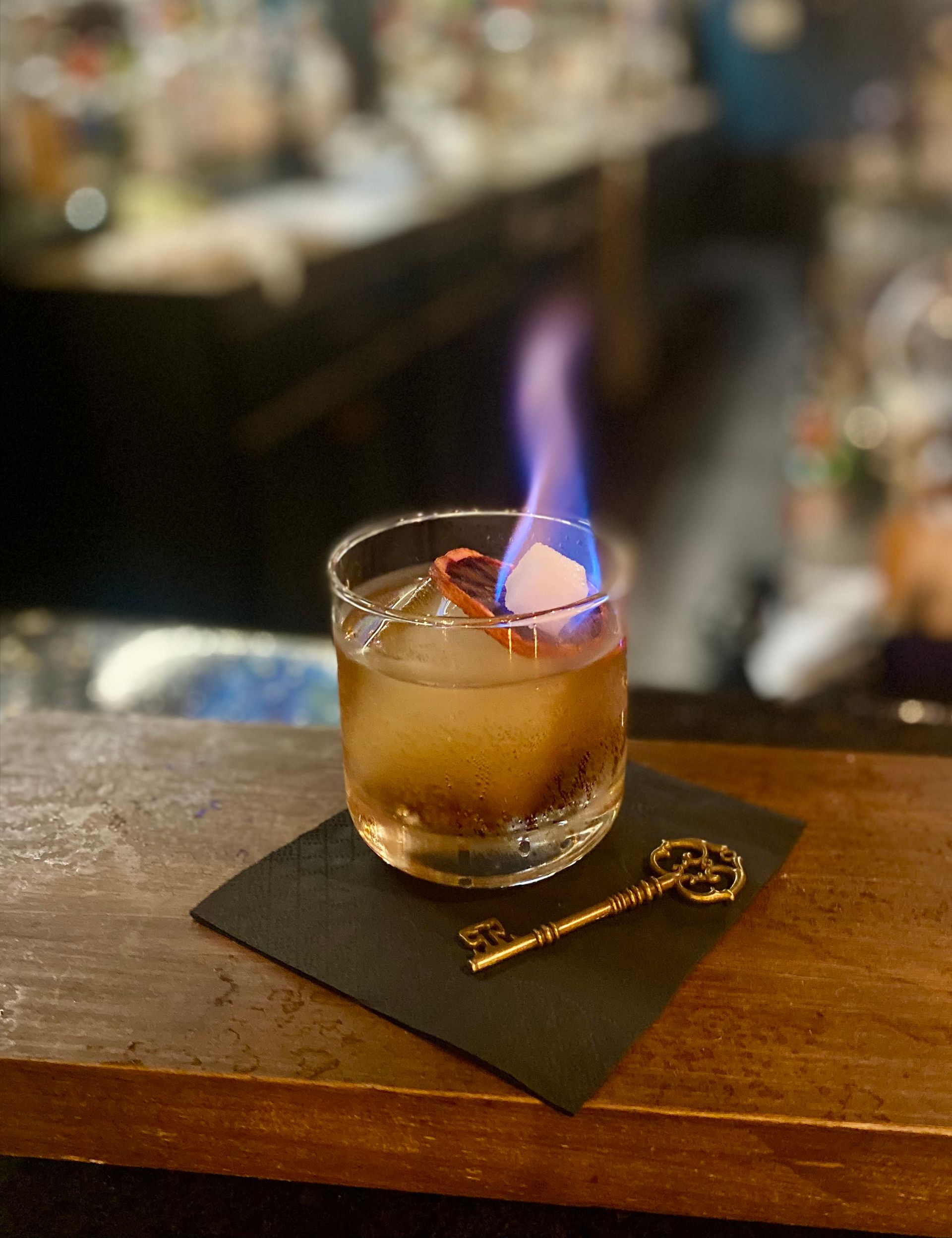 Flaming cocktail in a short glass on a bar counter beside a key-shaped stirrer