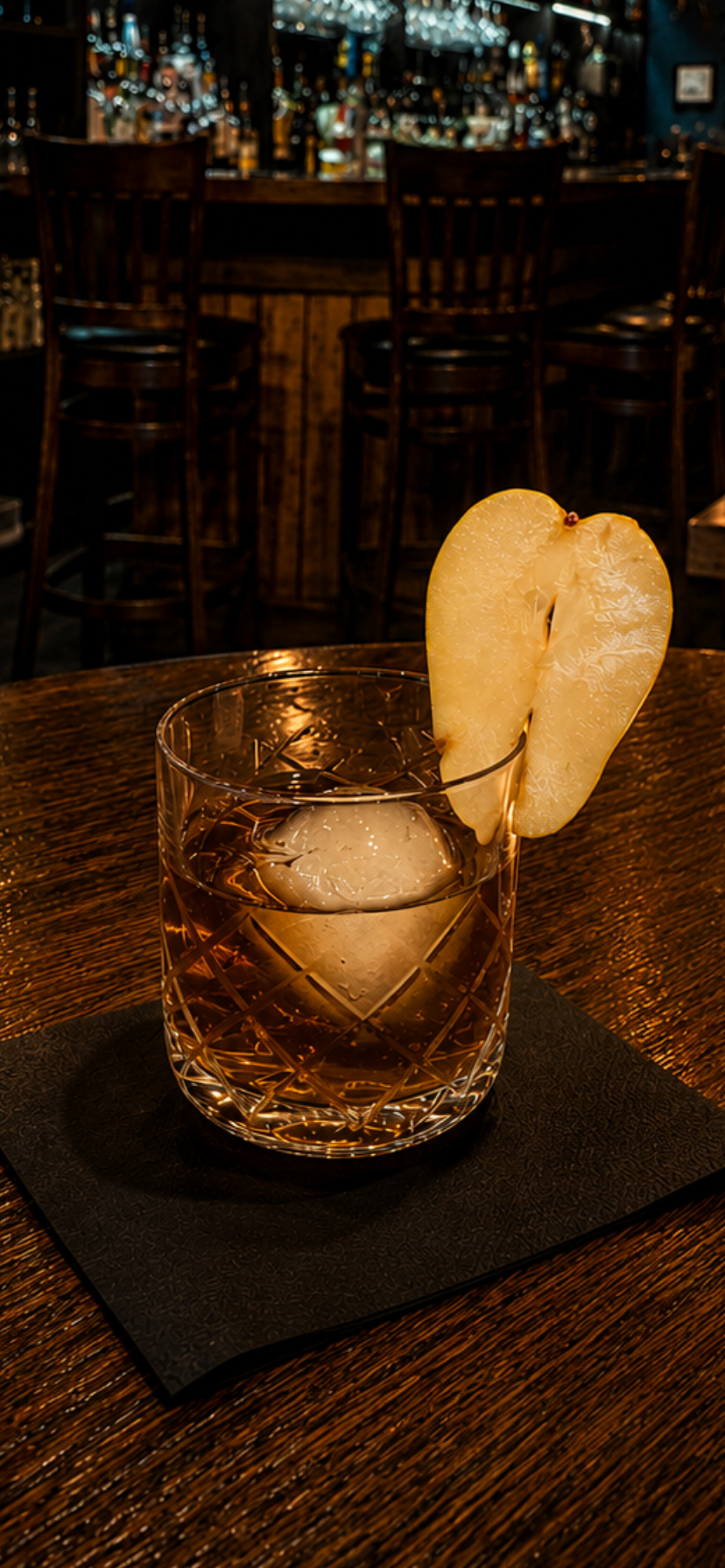 Whiskey glass with ice and pear garnish on a black coaster in a dimly lit bar