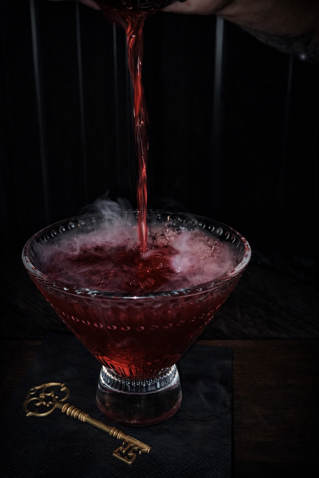 Dark red drink being poured into a coupe glass beside a gold key on a dark table.