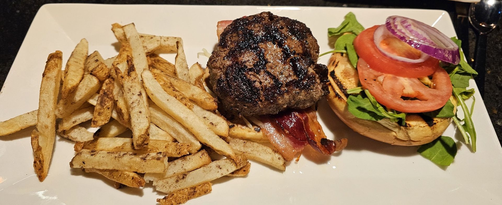Burger with fries on a white plate. Includes a bun, lettuce, tomato, red onion, bacon, and a grilled patty.