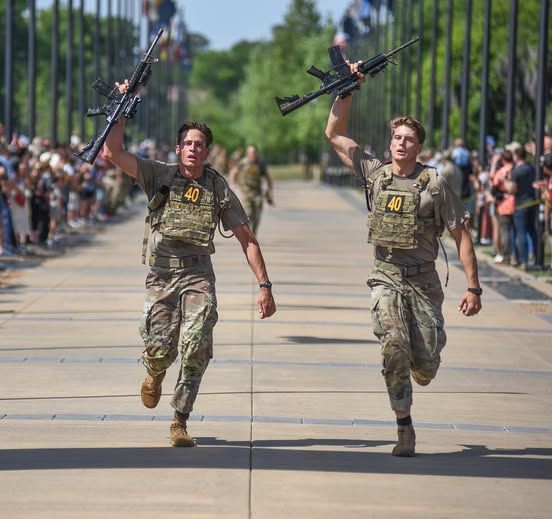 Two soldiers in camouflage, raising guns in victory as they run, crowds in background.