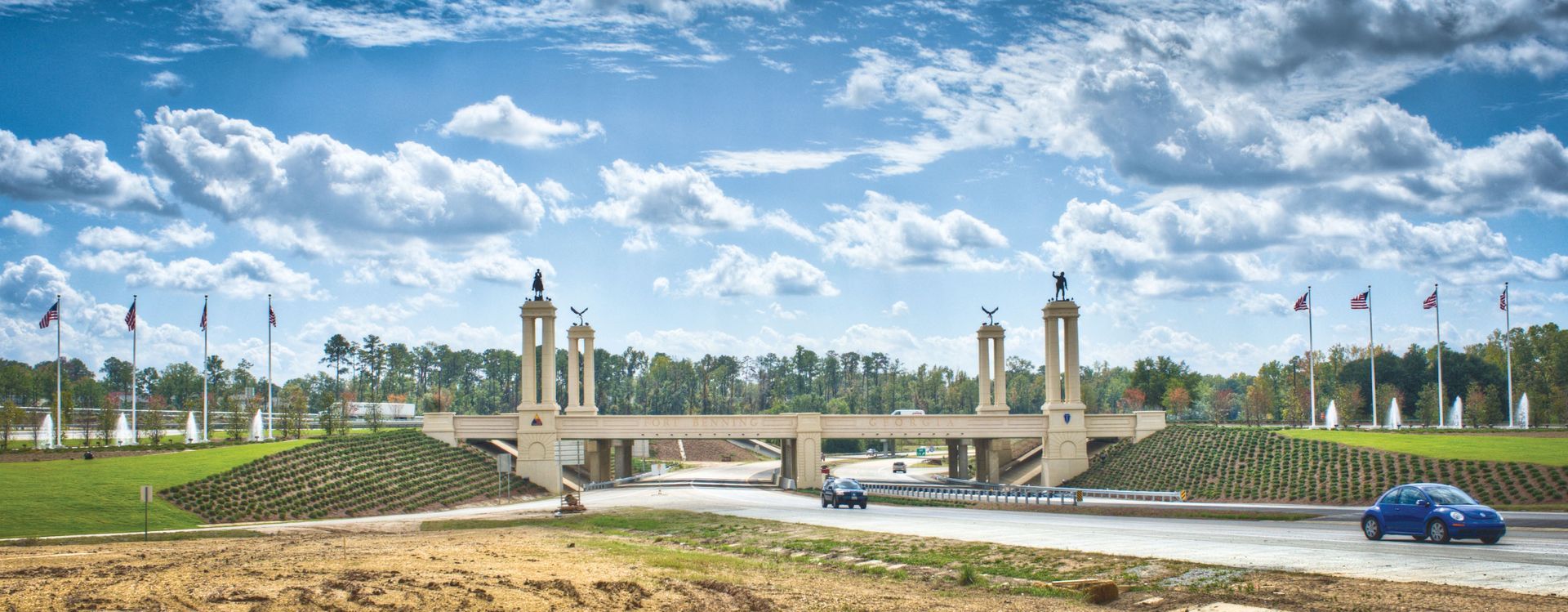 Bridge with towers, blue car, grassy hills, and cloudy sky.