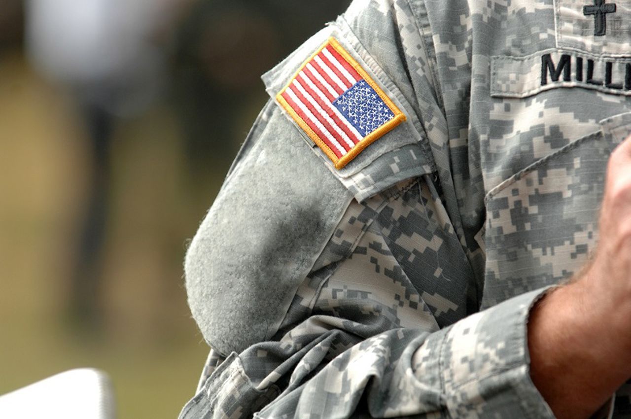 Close-up of U.S. Army uniform, featuring American flag patch on shoulder, name tag, and camouflage pattern.