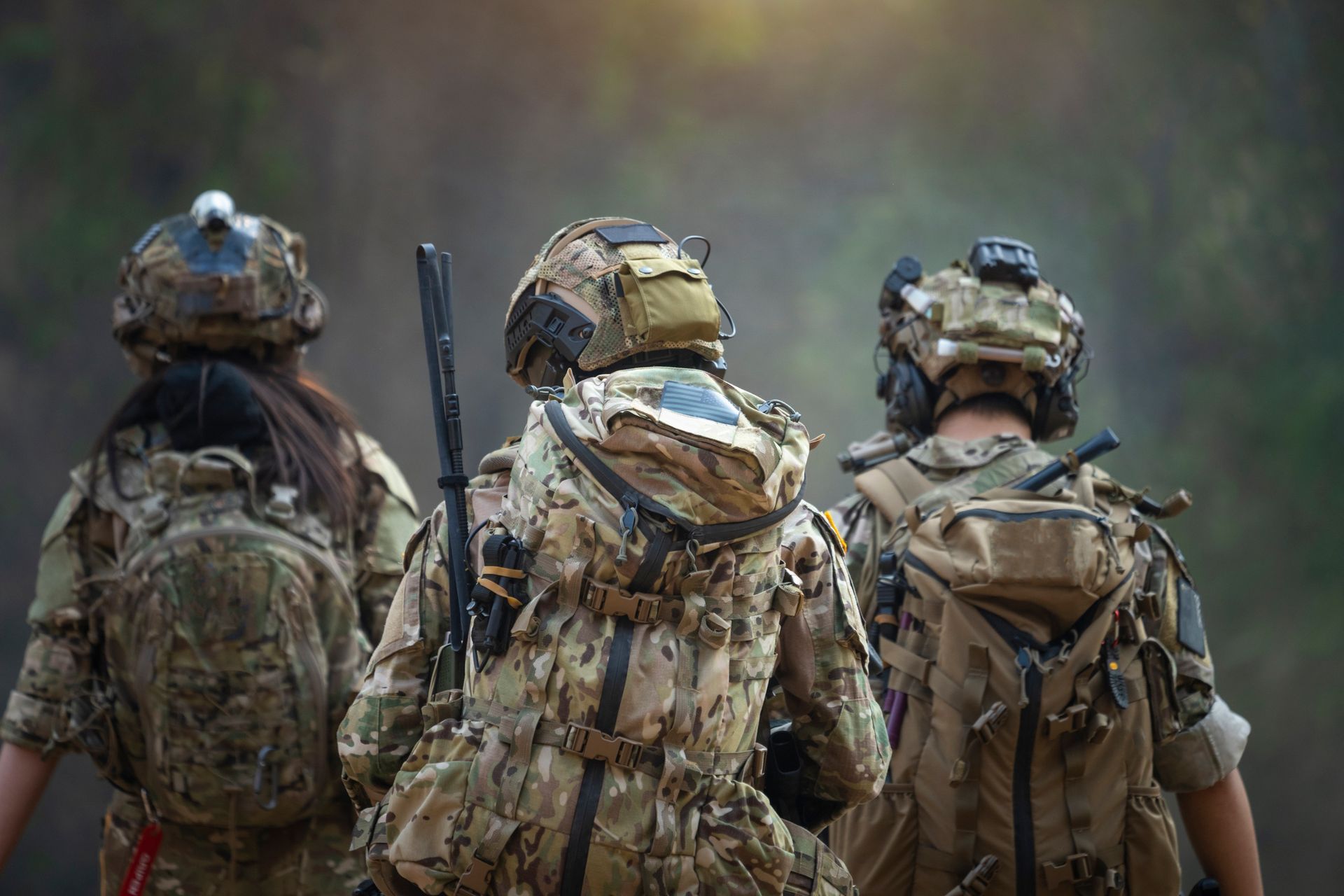 Three soldiers in camouflage gear walk through a forest, viewed from the back.