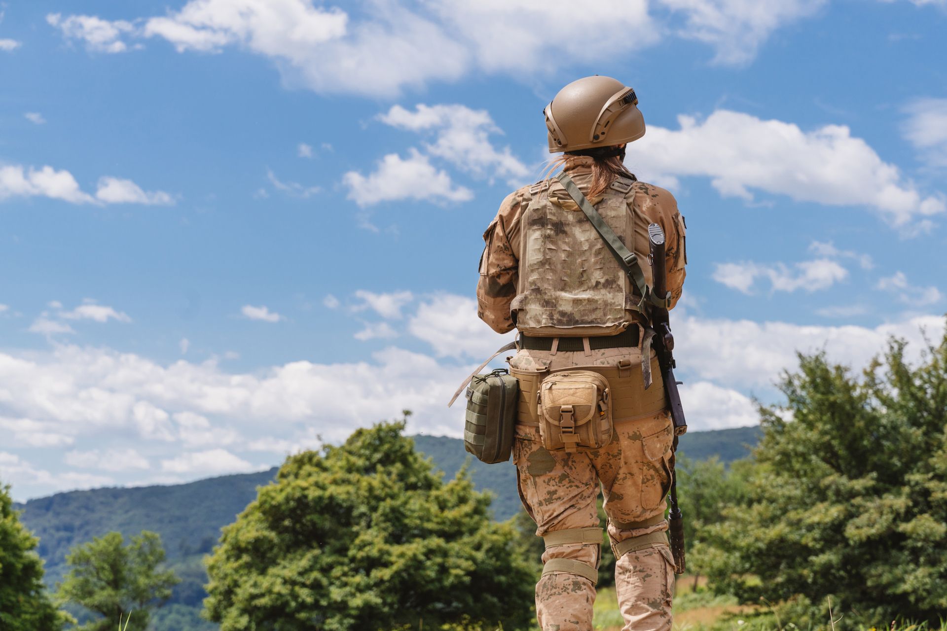 Soldier in desert camouflage gear gazes at the sky; mountains and trees in the background.