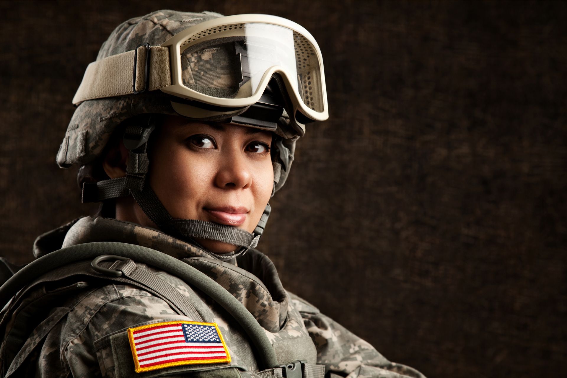 Female soldier in camouflage uniform, helmet, and goggles with an American flag patch.