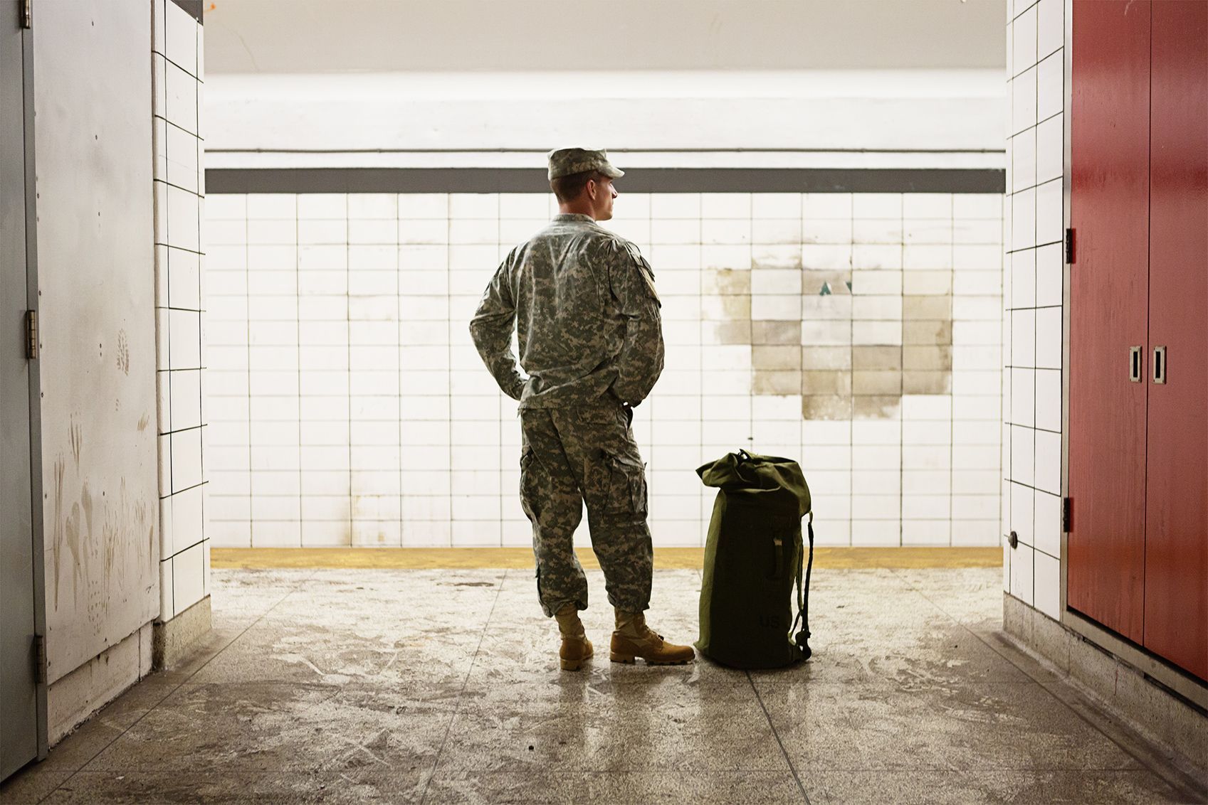 Soldier in camo uniform, standing with duffel bag in a subway station, looking out, hands in pockets.