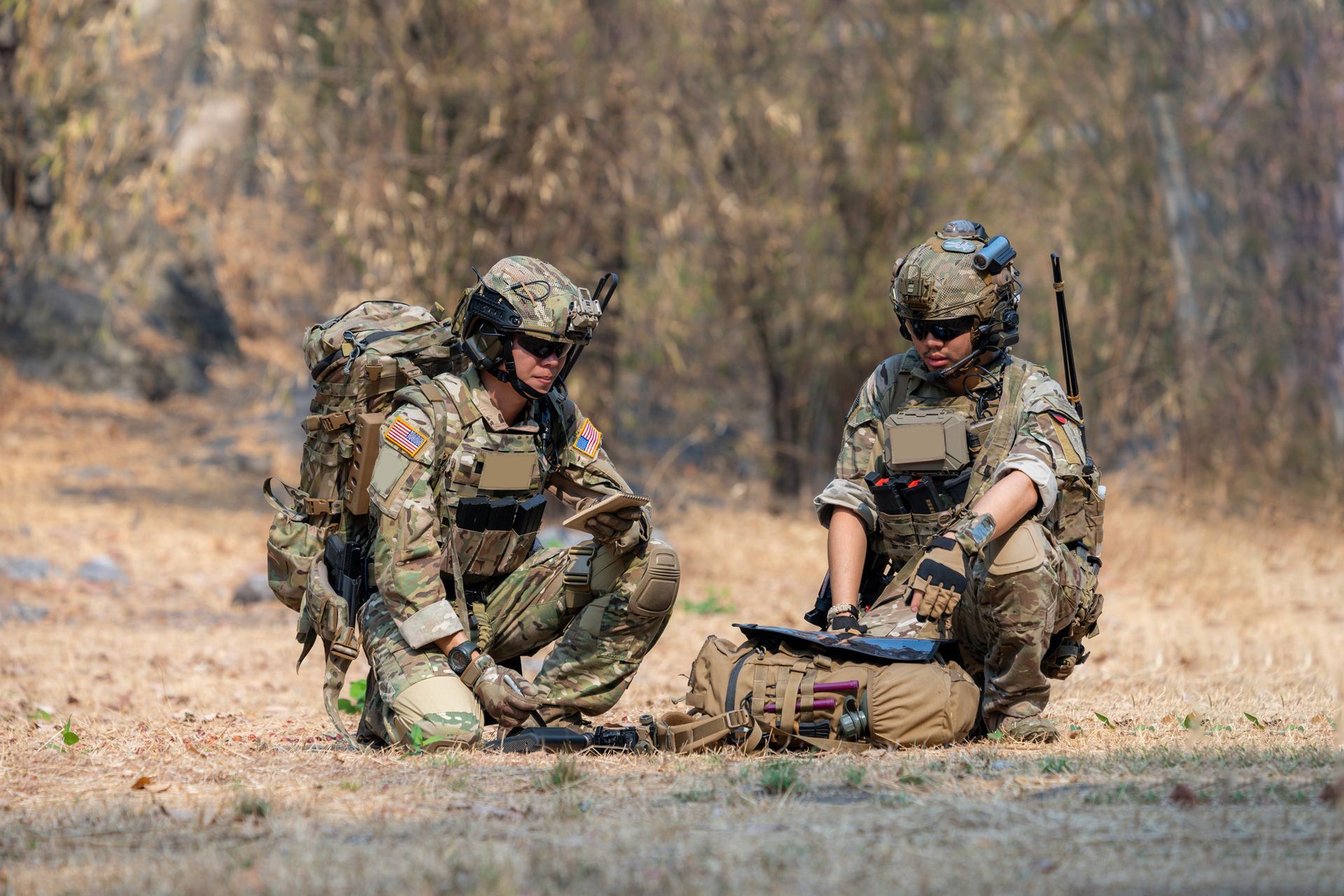 Two soldiers in camouflage gear crouch outdoors, examining equipment.