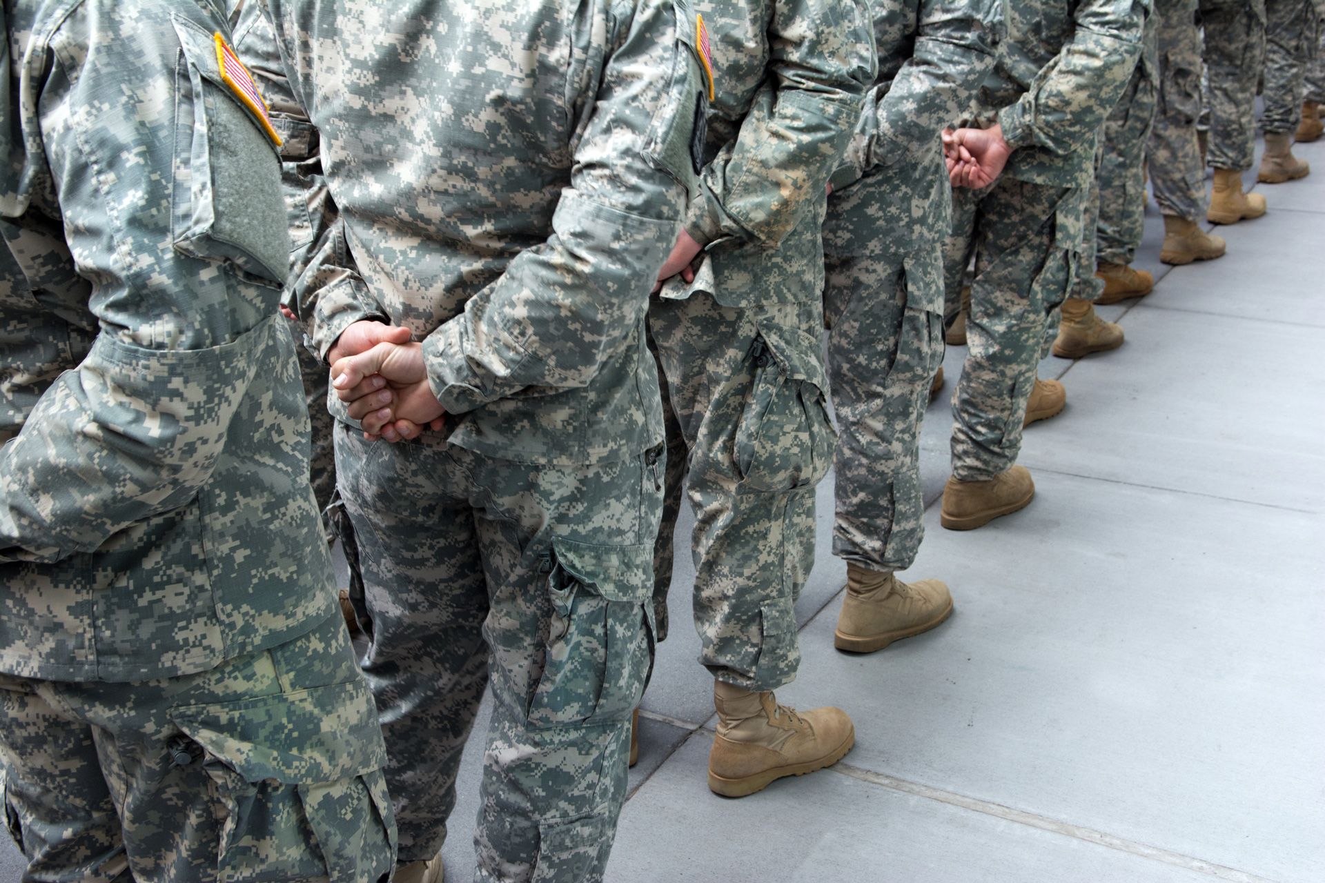 Soldiers in camouflage uniforms stand in line, hands clasped behind their backs.